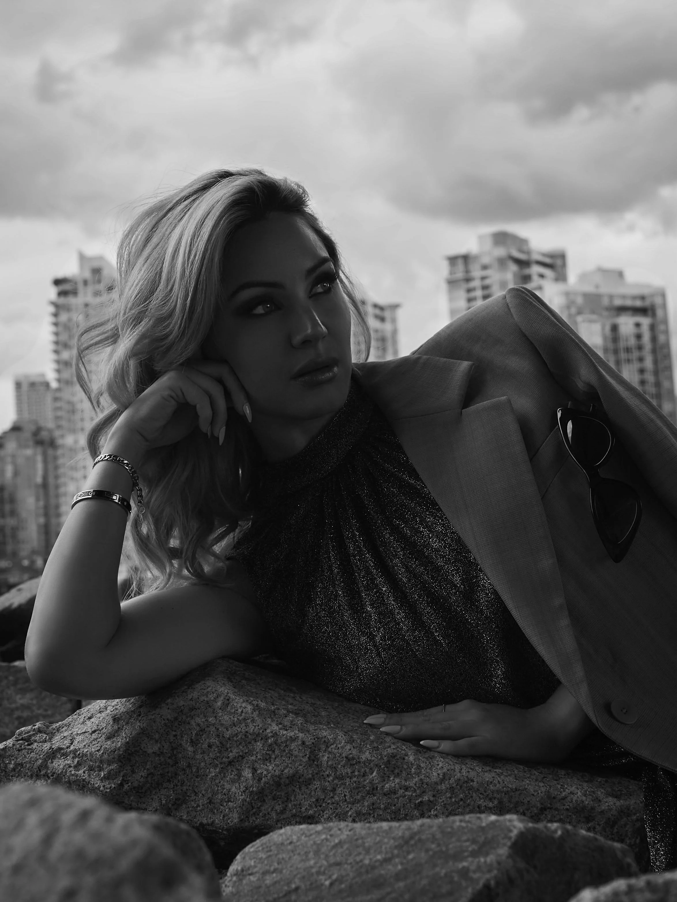 A woman with wavy blonde hair is lying on rocks outdoors, resting her head on her hand. She is wearing a blazer and a shimmery top, with sunglasses hanging from her blazer pocket. The background shows tall buildings and a cloudy sky.