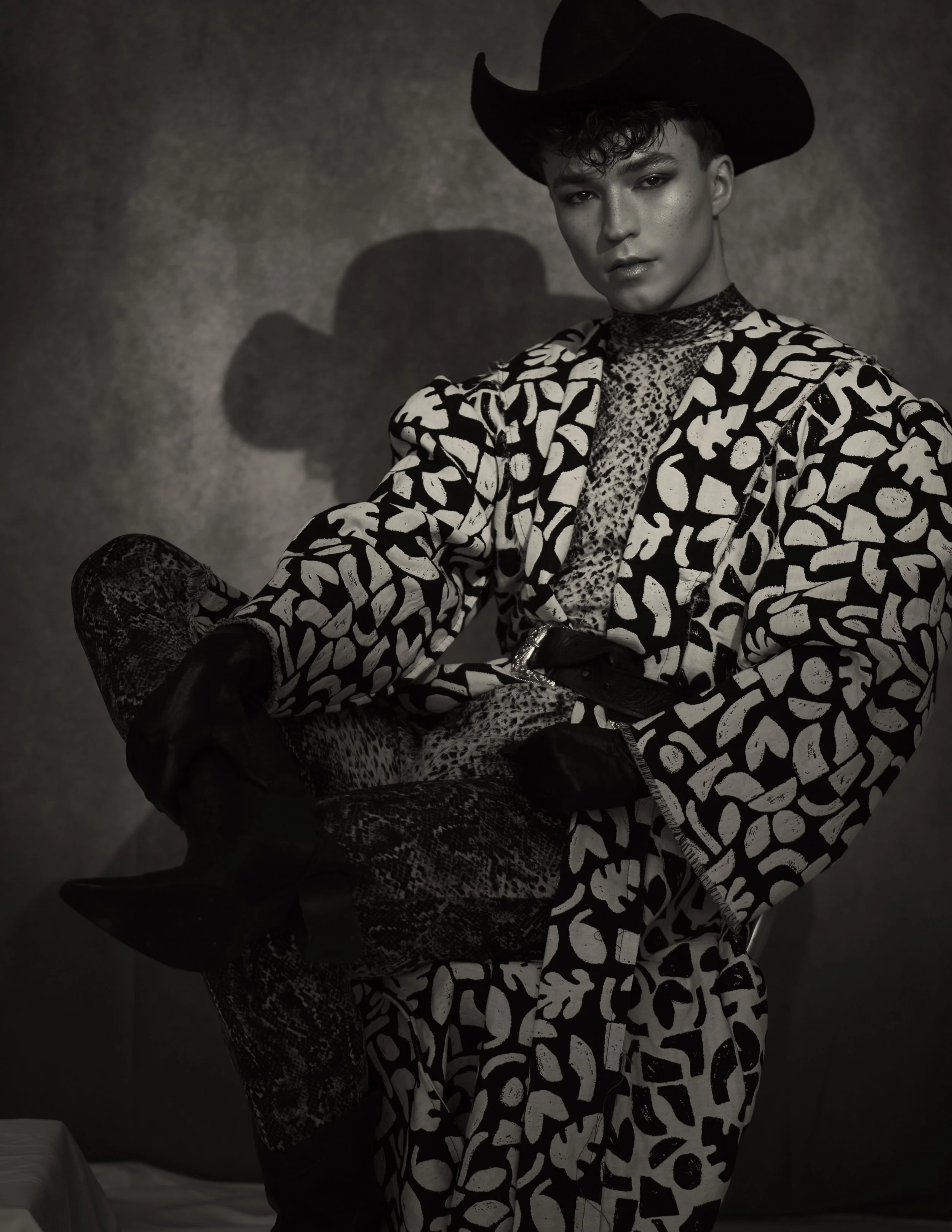 A woman in a patterned outfit with a wide-brimmed hat, sitting on a stool, in a black-and-white photo with a textured background.
