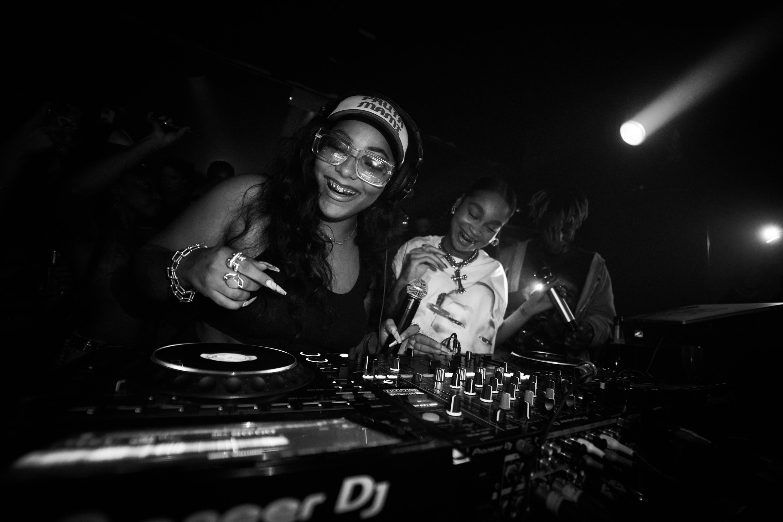 A black-and-white photo of two women DJing at a club, with a crowd in the background. The woman in the foreground is wearing large glasses, headphones, and smiling, pointing towards the camera. The second woman is smiling and holding a microphone.