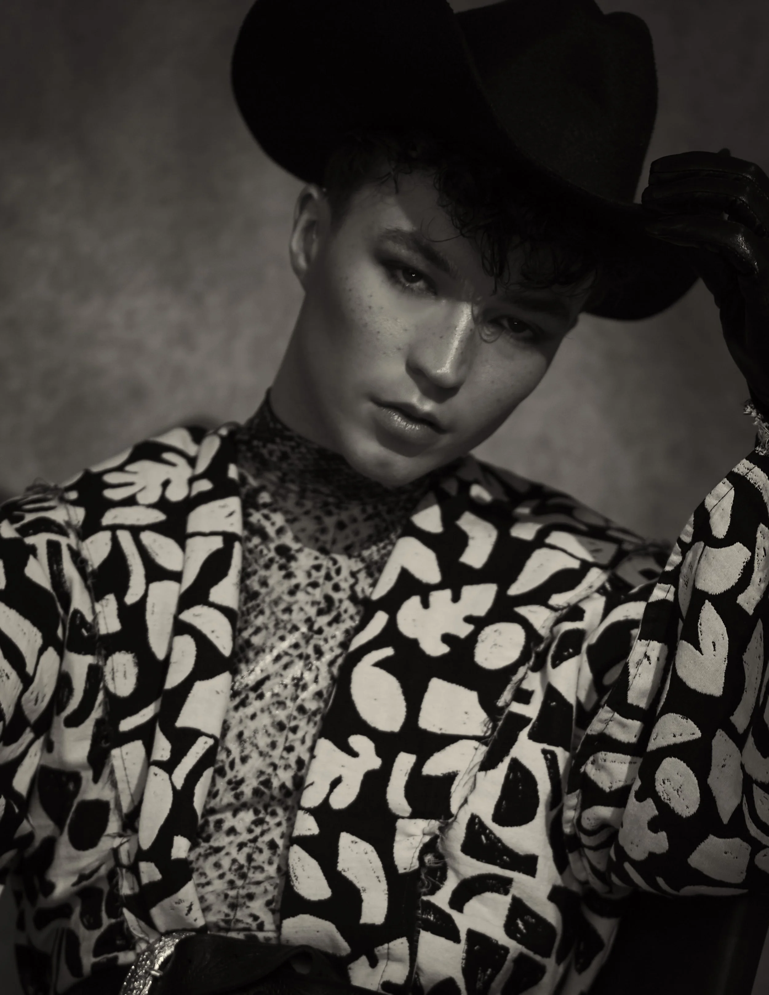 A black and white photo of a person wearing a wide-brimmed hat, leopard print top, and a patterned blazer with bold print, posing with a serious expression.