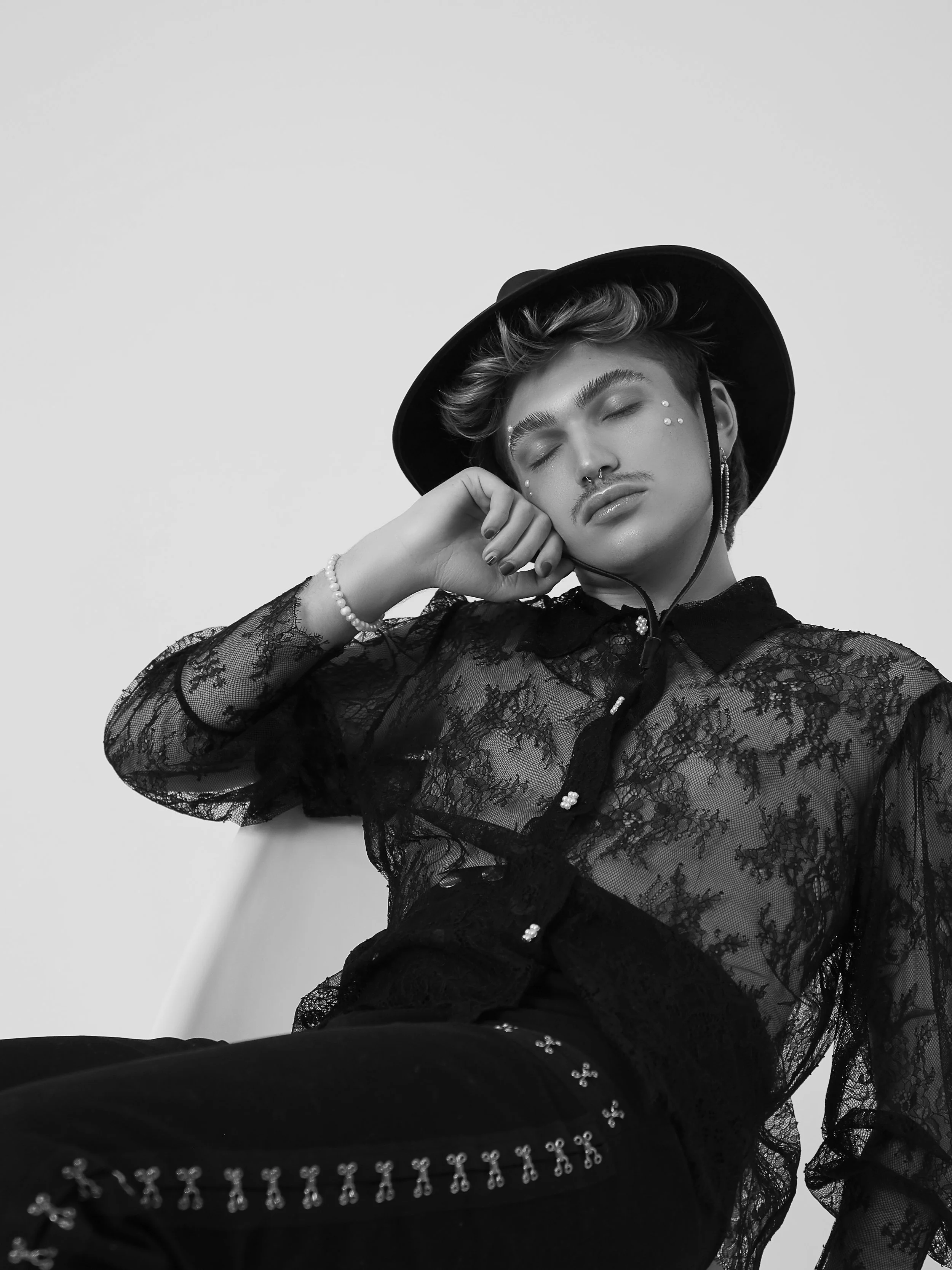 Black and white photo of a young person with short, styled hair, wearing a wide-brimmed hat, lace top, and jewelry, posing with closed eyes and a relaxed expression.
