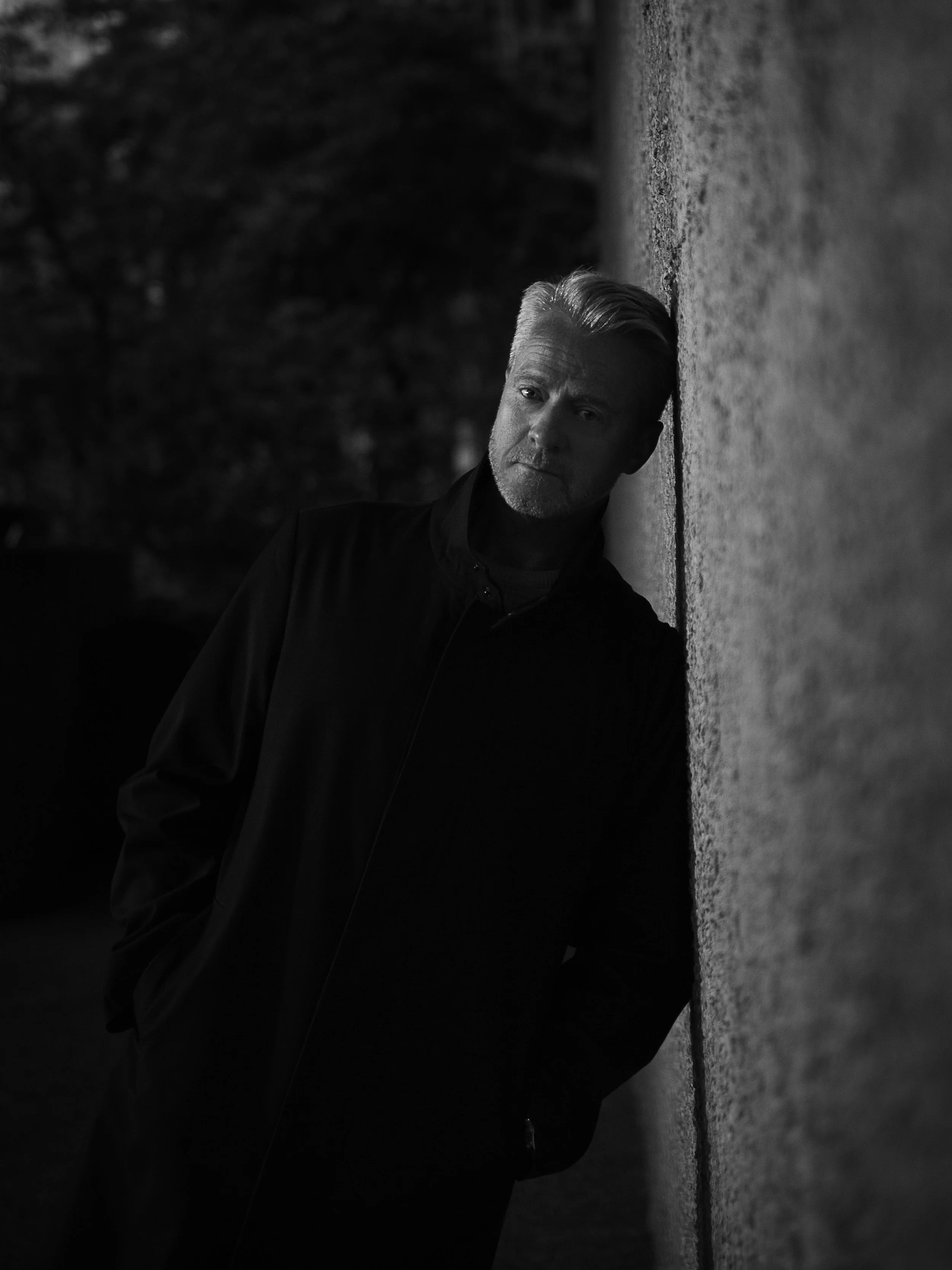 A black and white photo of a middle-aged man with gray hair and a beard, leaning against a wall outdoors at night, with a contemplative expression.
