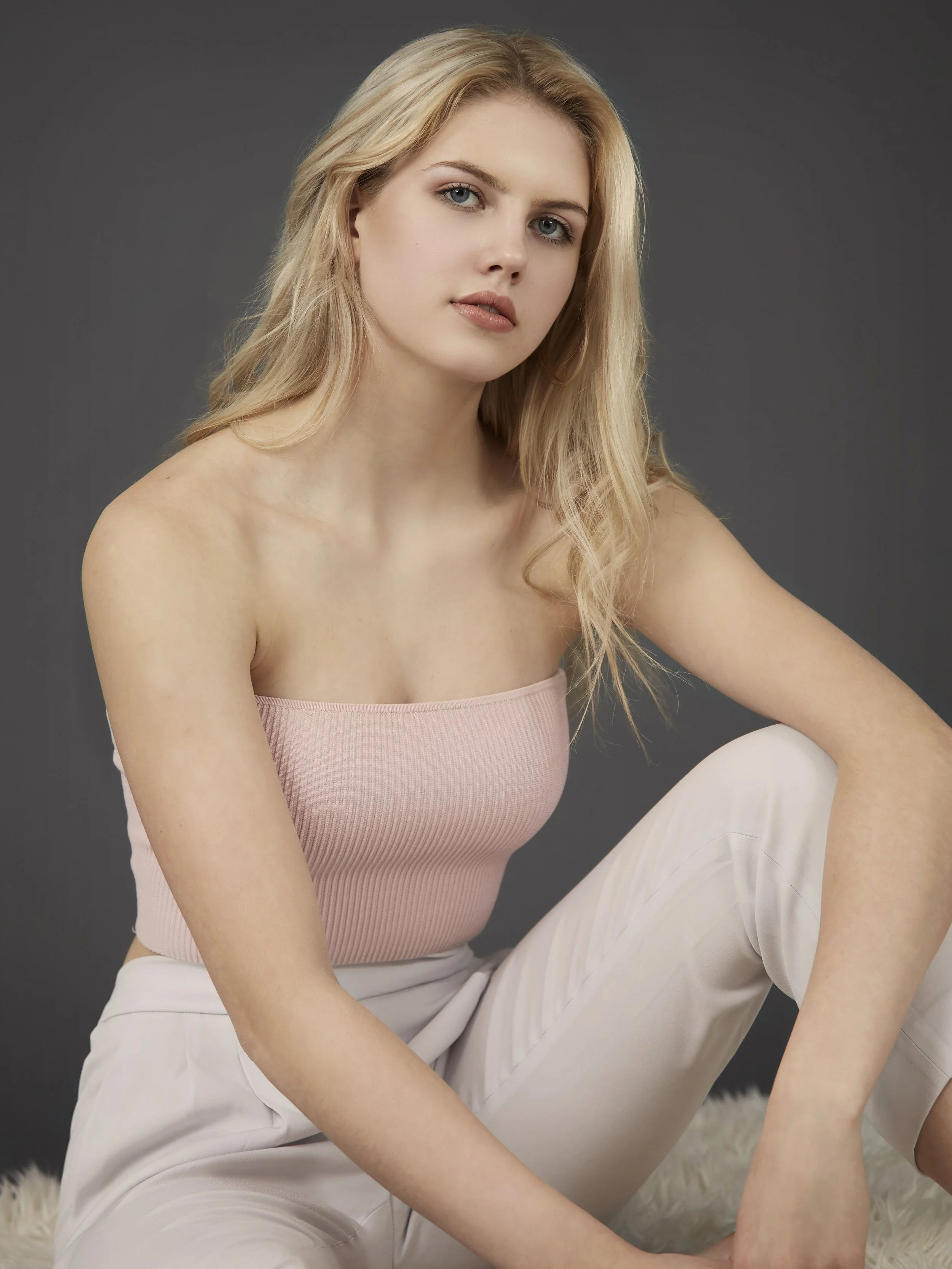 A young woman with long blonde hair, blue eyes, and natural makeup, sitting on a soft white rug against a dark gray background, wearing a strapless pink top and white pants.