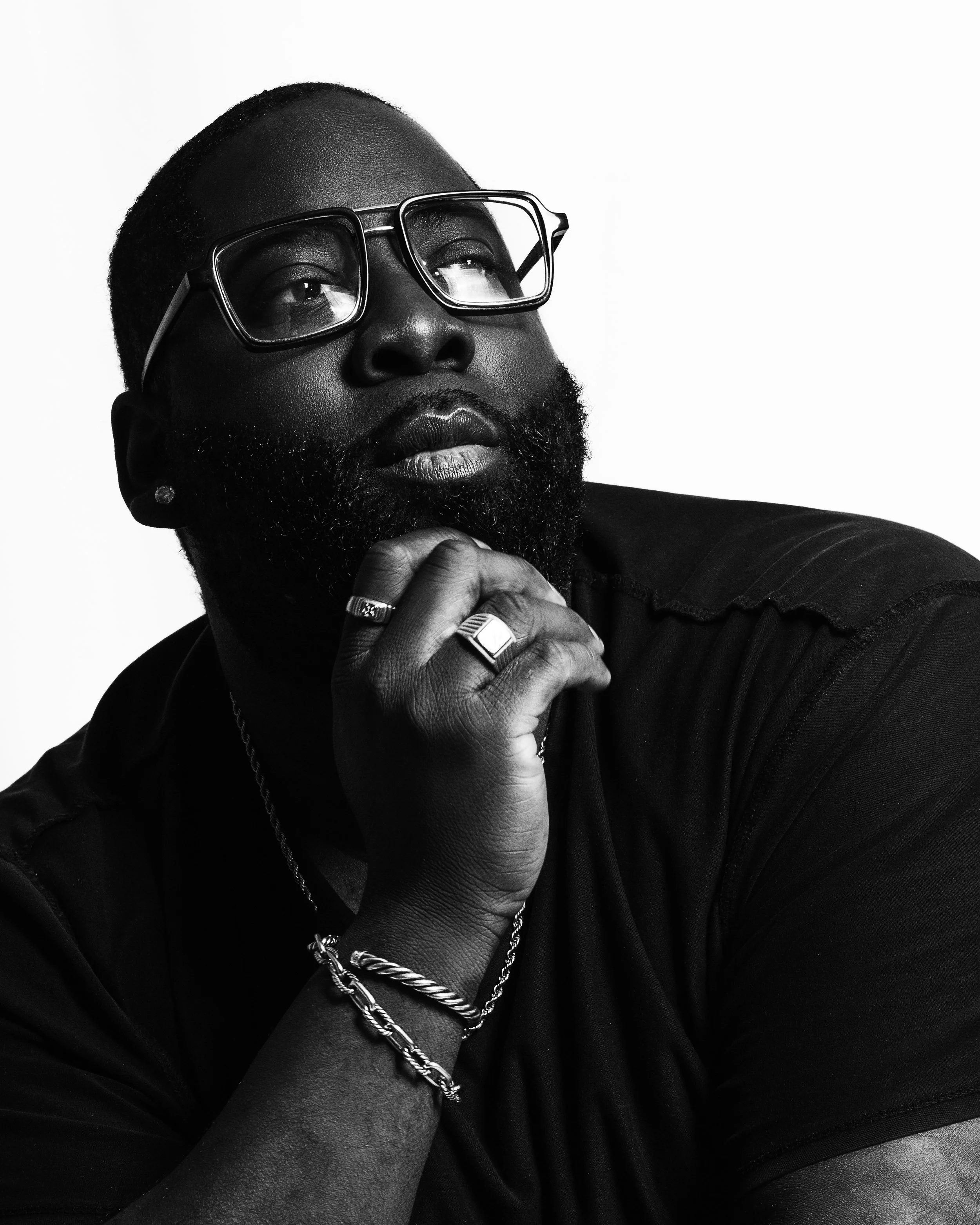 Black and white portrait of a man wearing glasses with hand on chin, jewelry including rings, bracelets, and chain, with a beard and short hair, looking contemplative.
