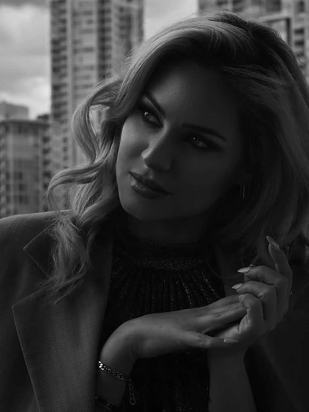A black and white photo of a woman with wavy hair, looking out a window with a cityscape background. She is wearing a blazer and has her hands clasped near her face.