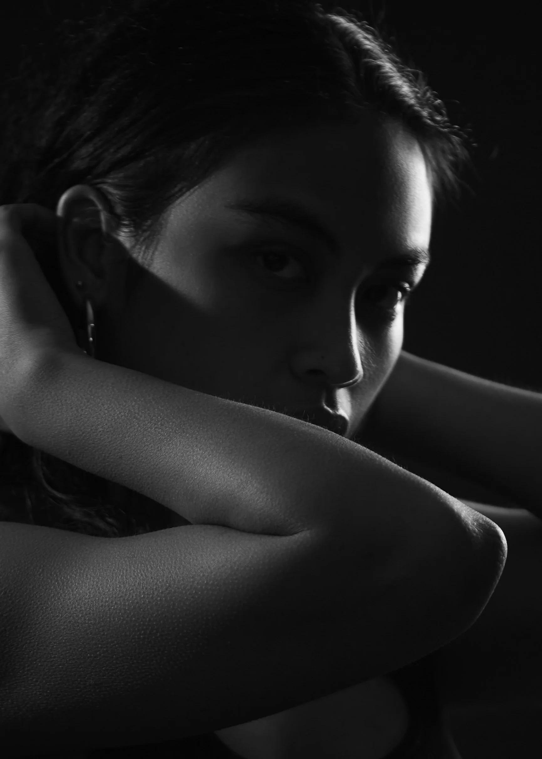 A black and white close-up photograph of a woman with her arms crossed, partially covering her face, with dramatic lighting highlighting her features and hair.