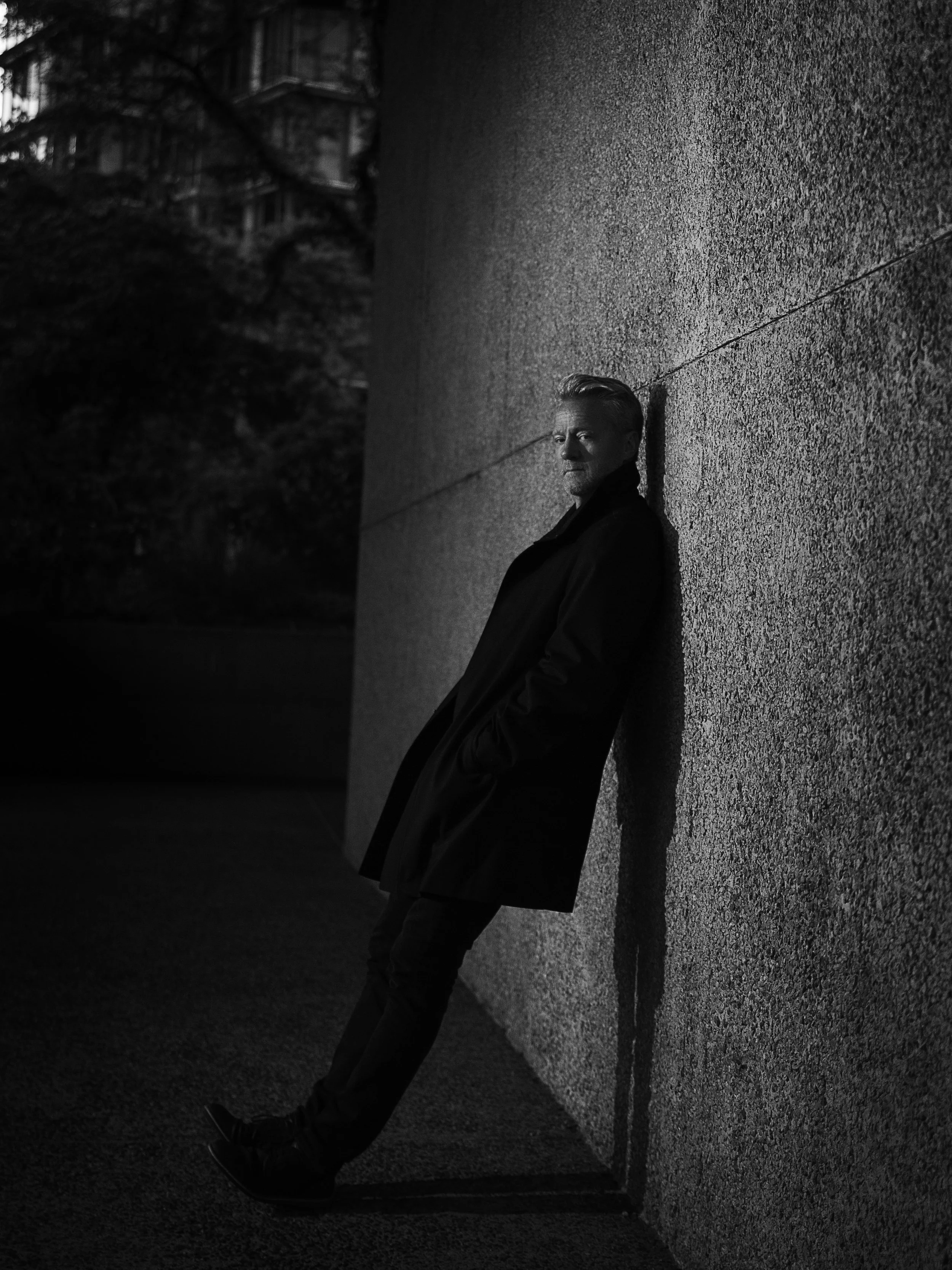A man leaning against a textured wall with his hands in his pockets, looking at the camera during nighttime or in low light.