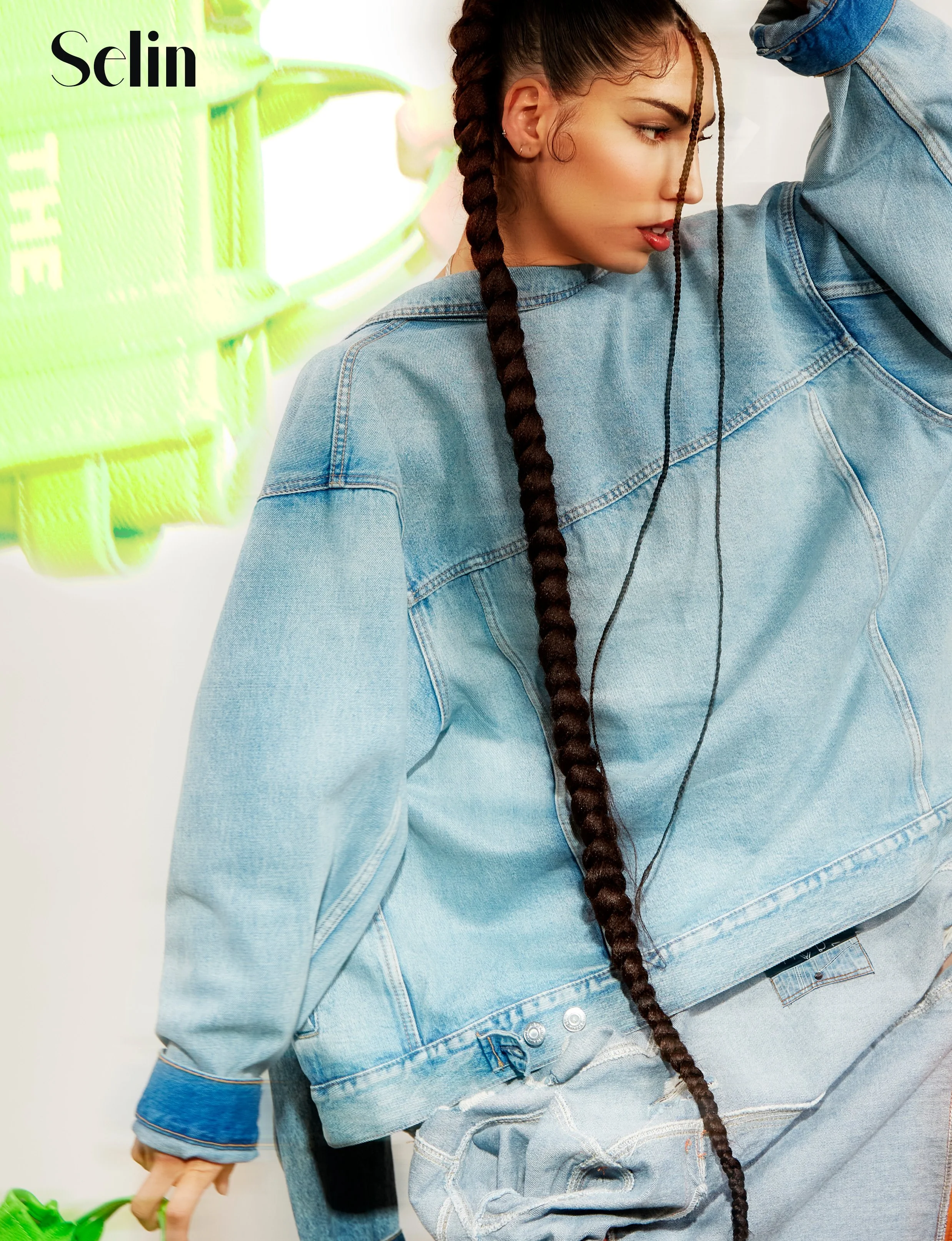 A woman with long braided hair in a denim jacket, holding a green leash, standing near a neon green sign with the word 'Selin' on it.