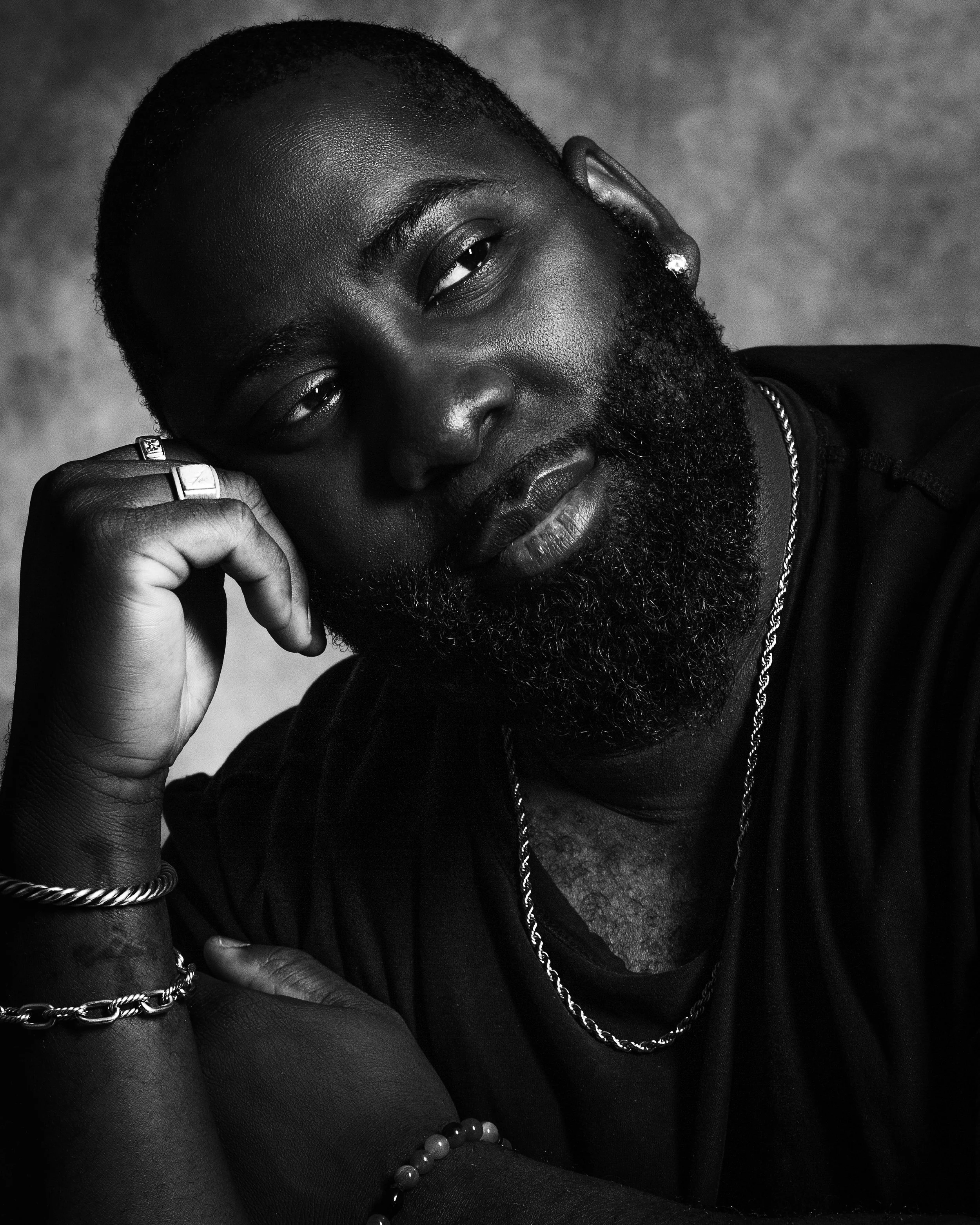 Black and white portrait of a man with a beard, short haircut, earrings, and layered necklaces, resting his head on his hand.