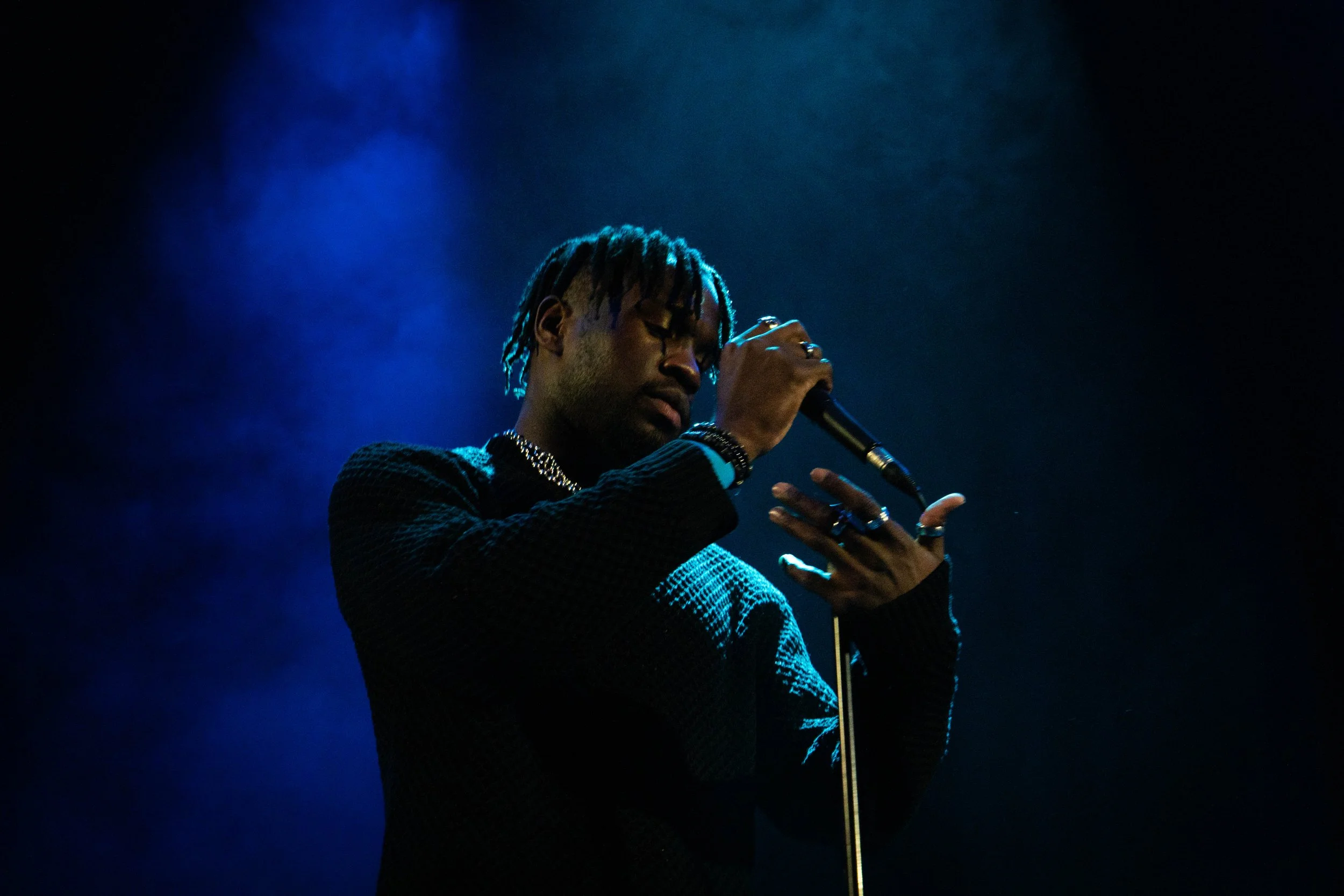 A man with dreadlocks singing into a microphone on stage, illuminated by blue stage lighting.