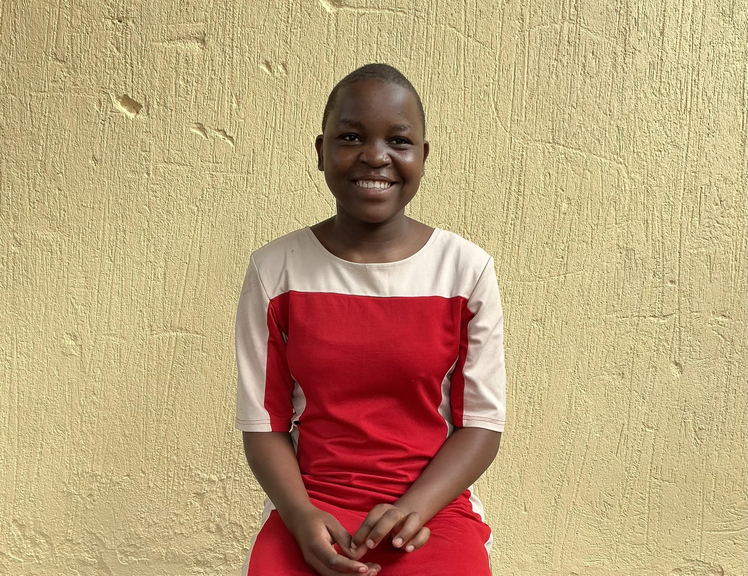 A young woman smiling, seated against a yellow textured wall, wearing a red and beige dress with three-quarter sleeves.