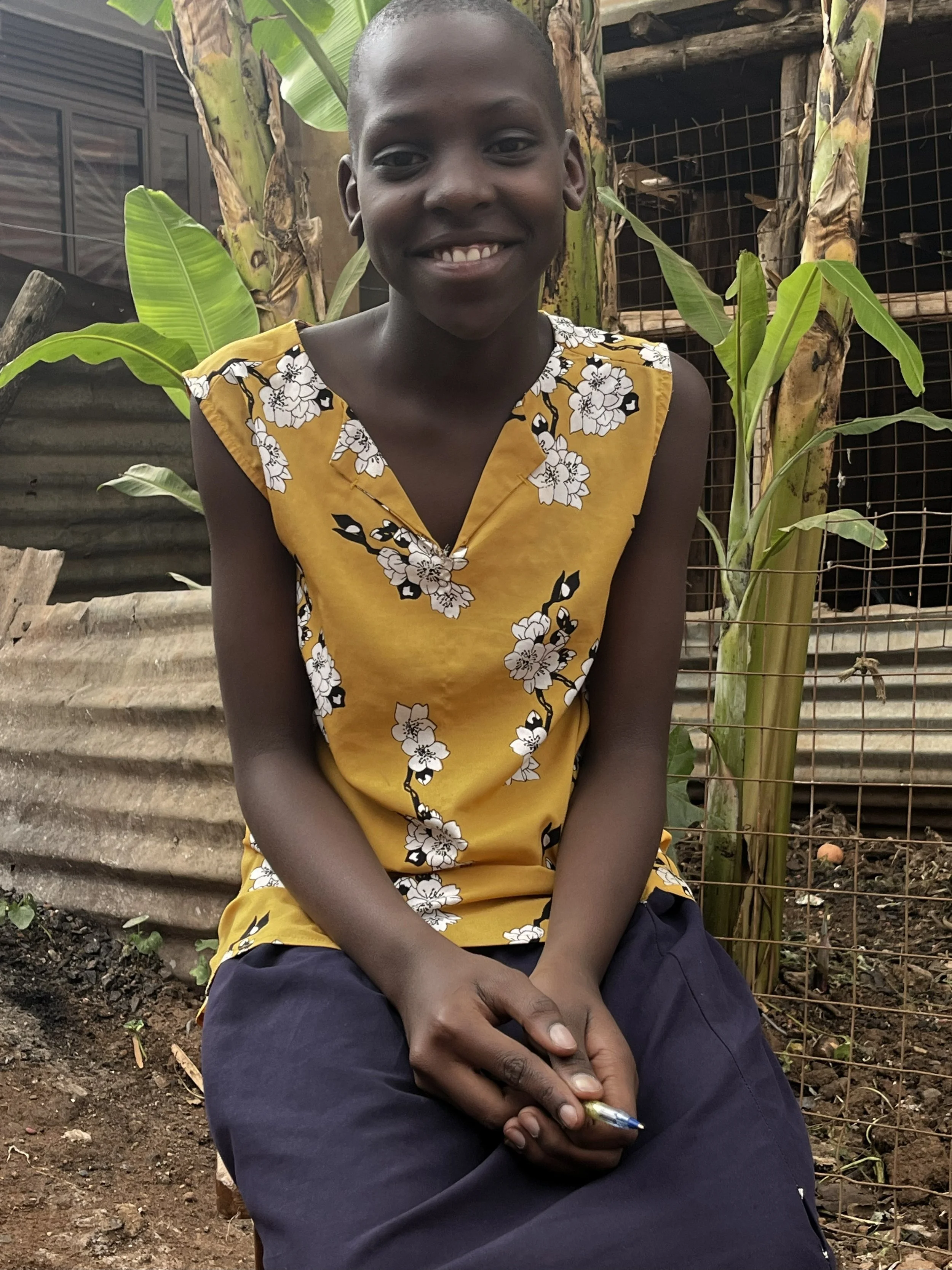 A smiling young boy with a shaved head, wearing a sleeveless yellow top with white and black floral patterns, sitting outdoors in front of greenery and a wire fence.