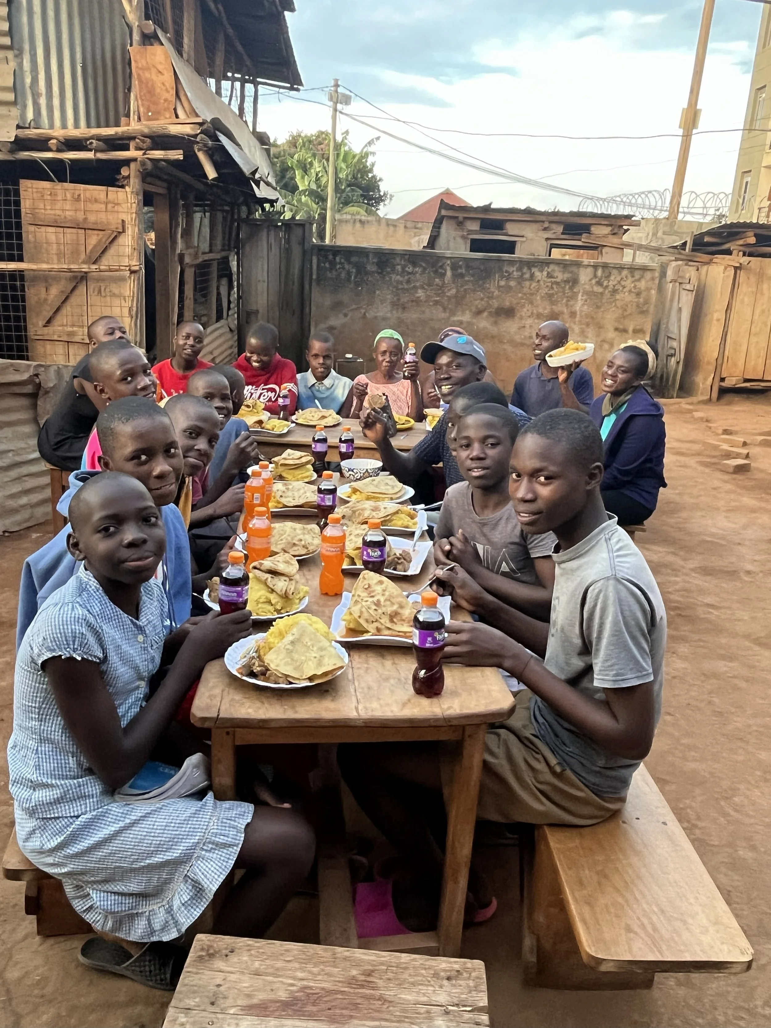 A group of children and a few adults are sitting around a long wooden table outdoors, enjoying a meal together with plates of food, bottles of juice, and some snacks. The setting appears to be in a rustic, open area with makeshift structures and a dirt ground.