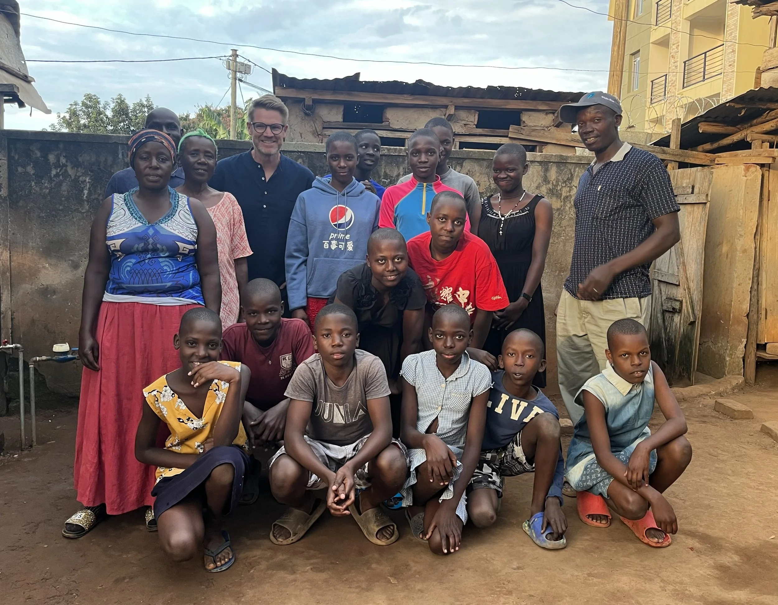 A diverse group of children and adults posing together in an outdoor setting with a wall and buildings in the background.