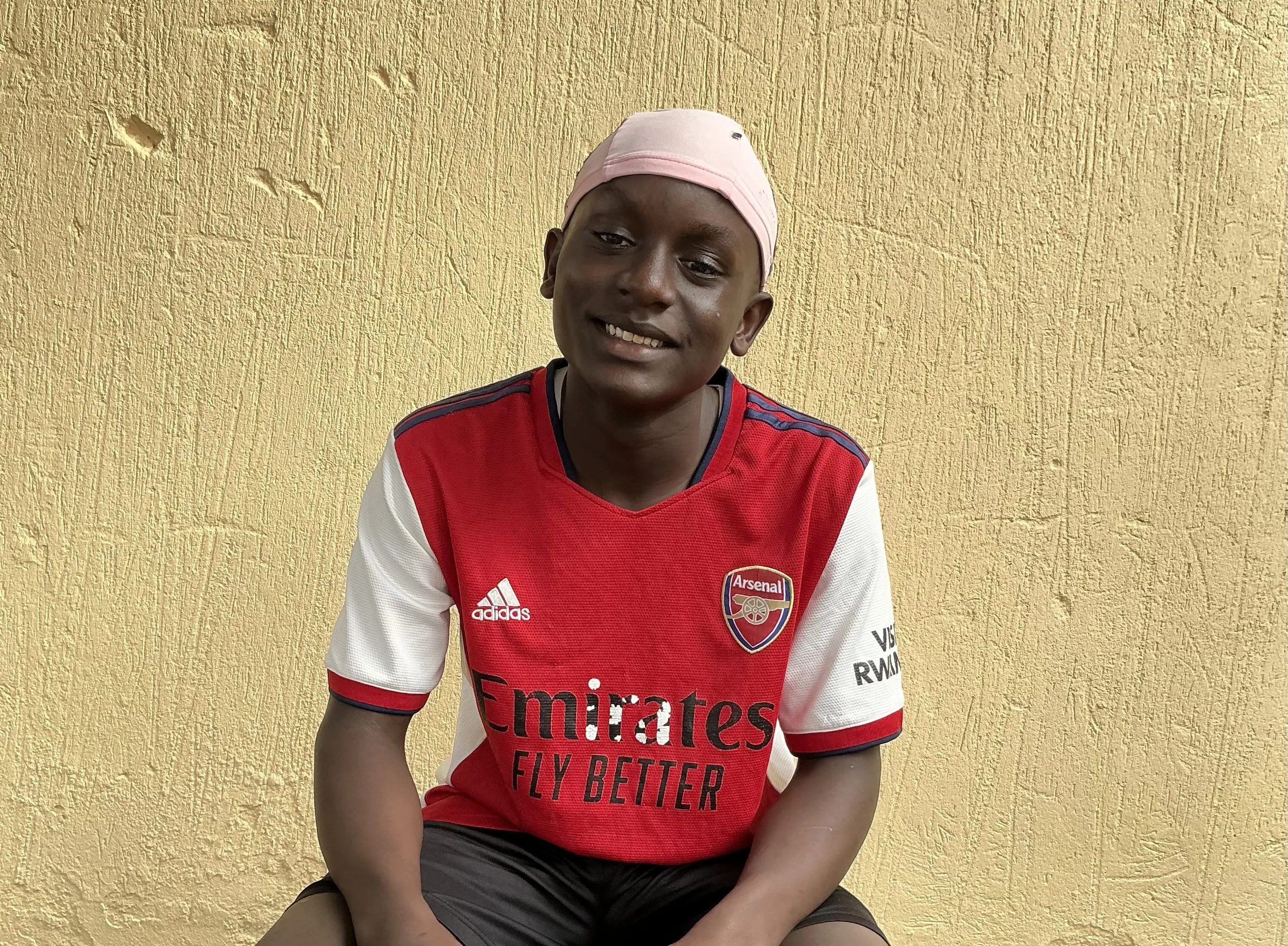 A young boy sitting against a textured yellow wall, wearing a red and white Arsenal football jersey and a pink head covering, smiling at the camera.
