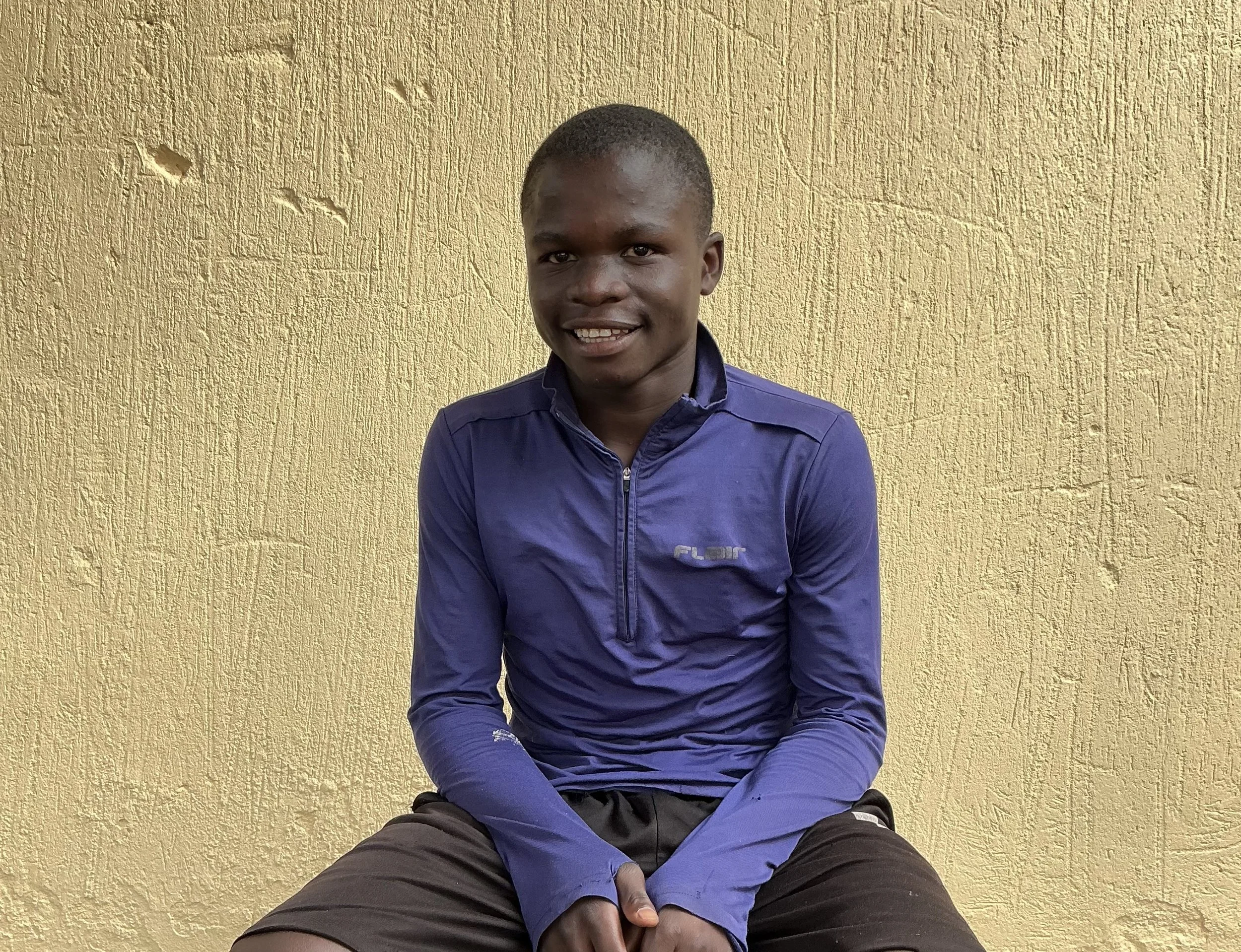 A young man in a blue jacket sitting against a yellow wall, smiling at the camera.