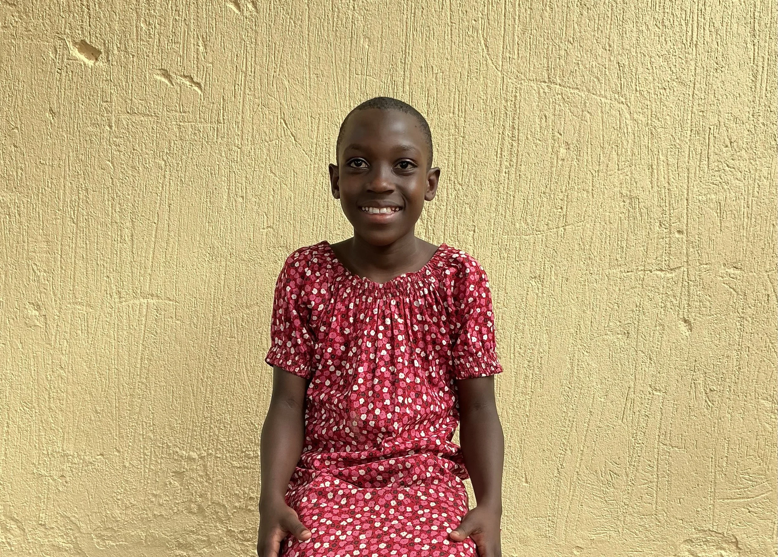 A young girl with a short haircut, smiling and sitting against a yellow textured wall. She is wearing a red dress with pink and white floral patterns.