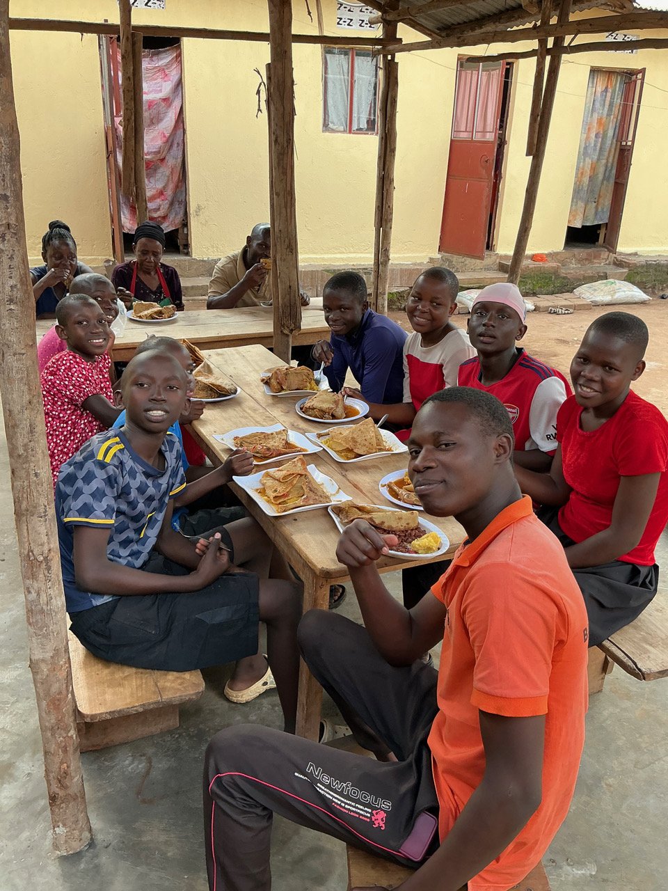 Group of children and a few adults sitting at wooden tables under a simple wooden structure, enjoying a meal with plates of food, in a modest outdoor setting.