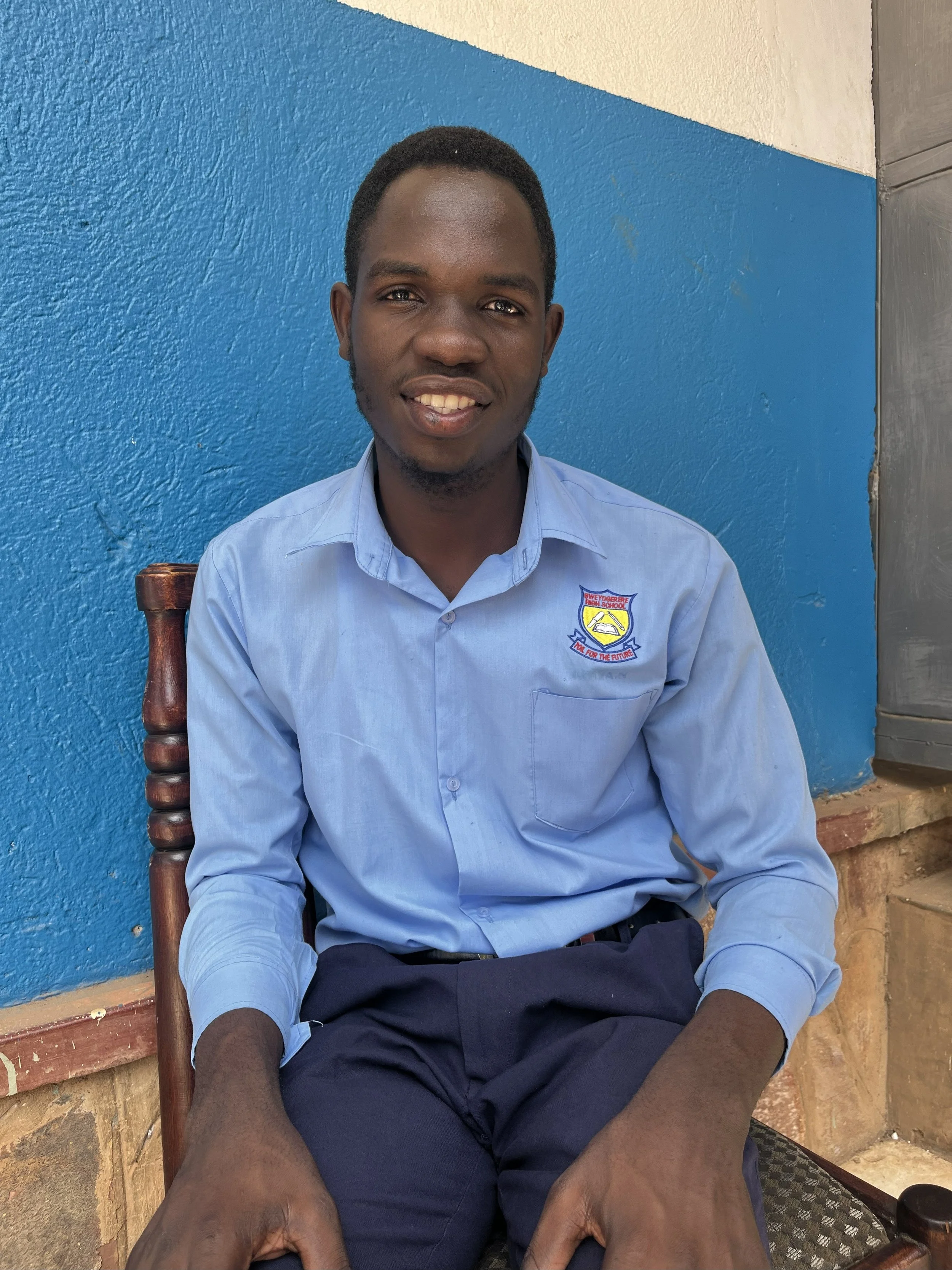 A young man with short black hair, smiling, seated against a blue wall. He is wearing a light blue shirt with a patch on the chest, dark pants, and sitting on a wooden chair.