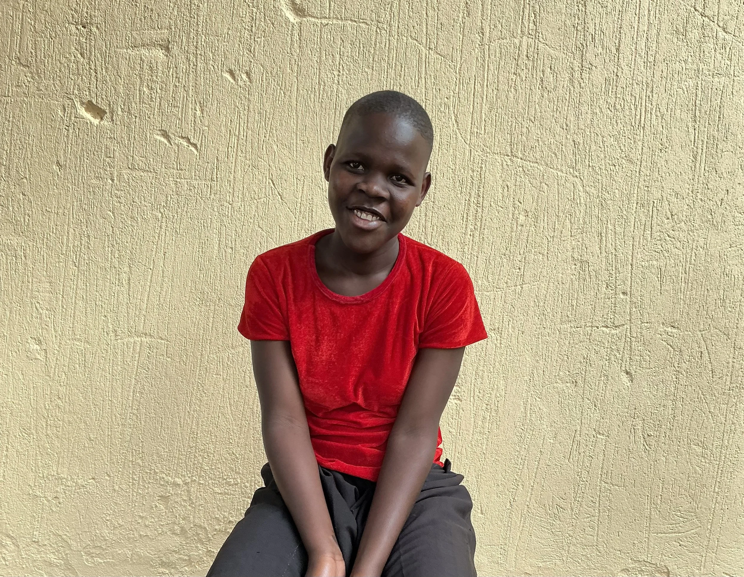 A young boy with short hair wearing a red t-shirt and dark pants, sitting in front of a textured yellow wall, smiling at the camera.