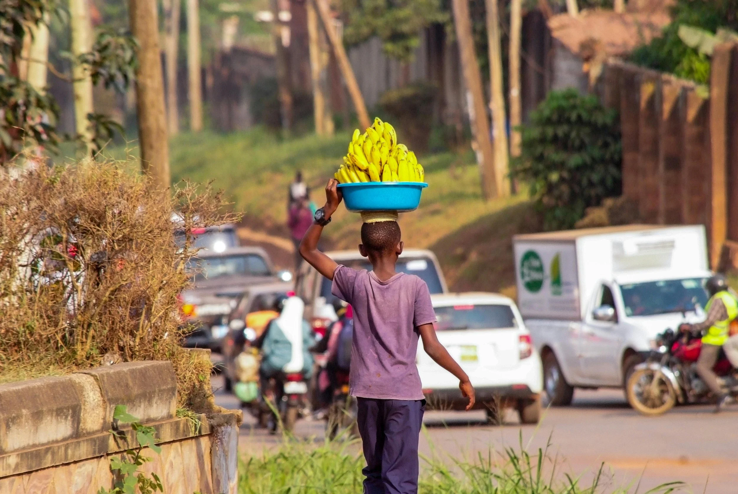 A young boy walking along a busy street carrying a large blue tray filled with bananas on his head.