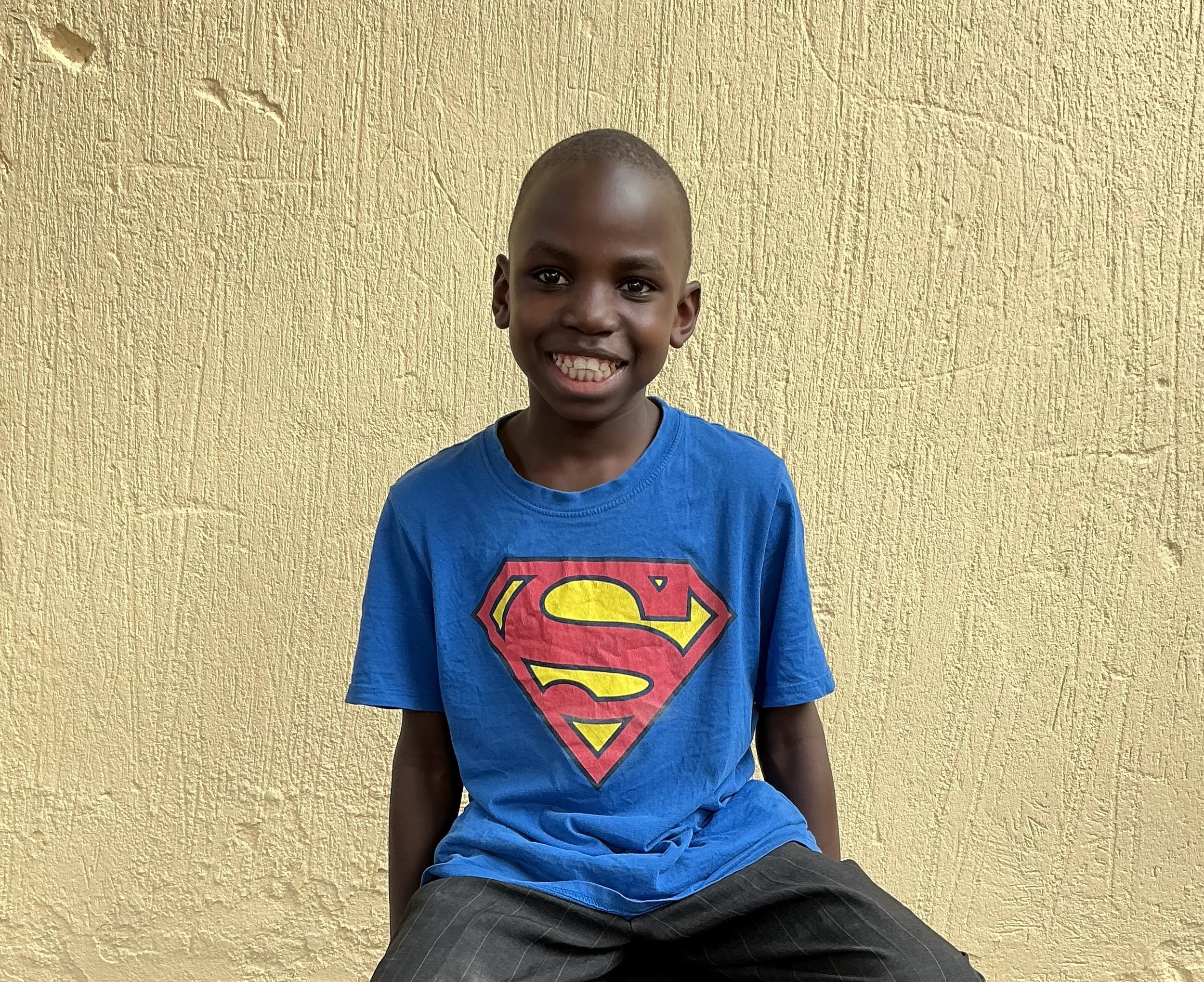 Young boy sitting against a yellow textured wall, smiling, wearing a blue Superman t-shirt and dark pants.