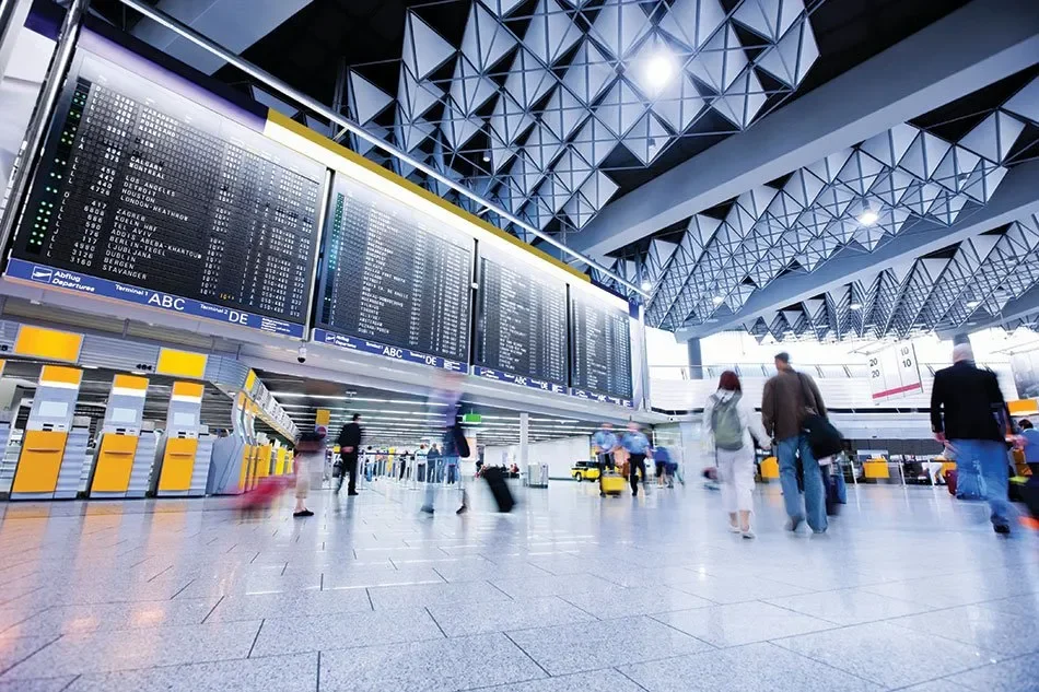 Passengers walking in an airport terminal, with flight information displayed on large digital screens overhead. The modern space features a geometric ceiling design and check-in counters with yellow accents.