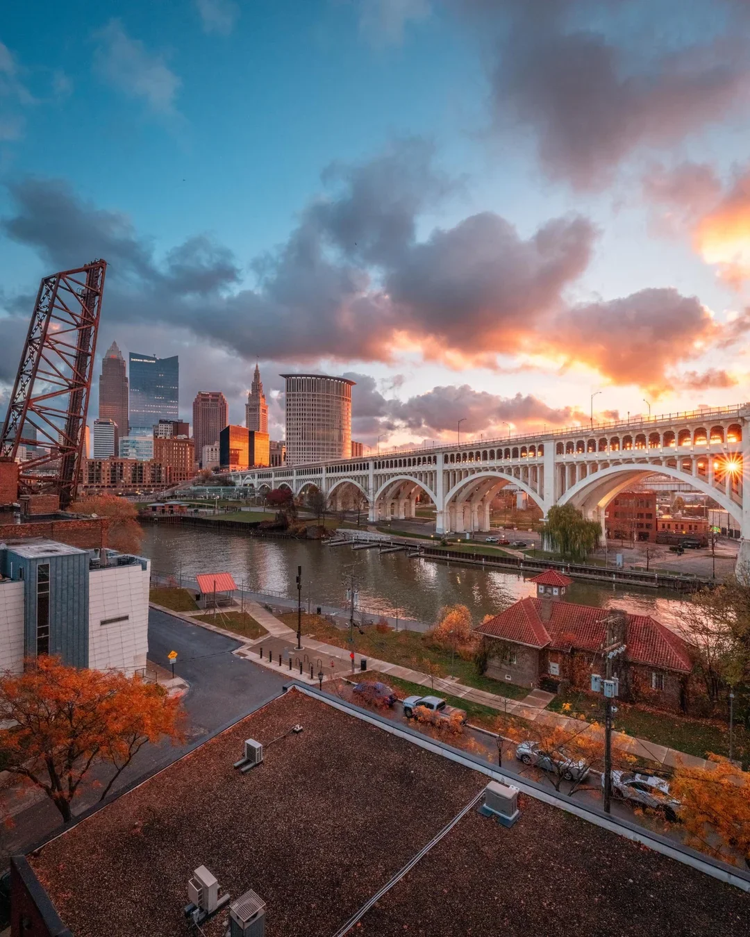 City skyline at sunset with tall buildings, a bridge over a river, and a sky with clouds and the setting sun.