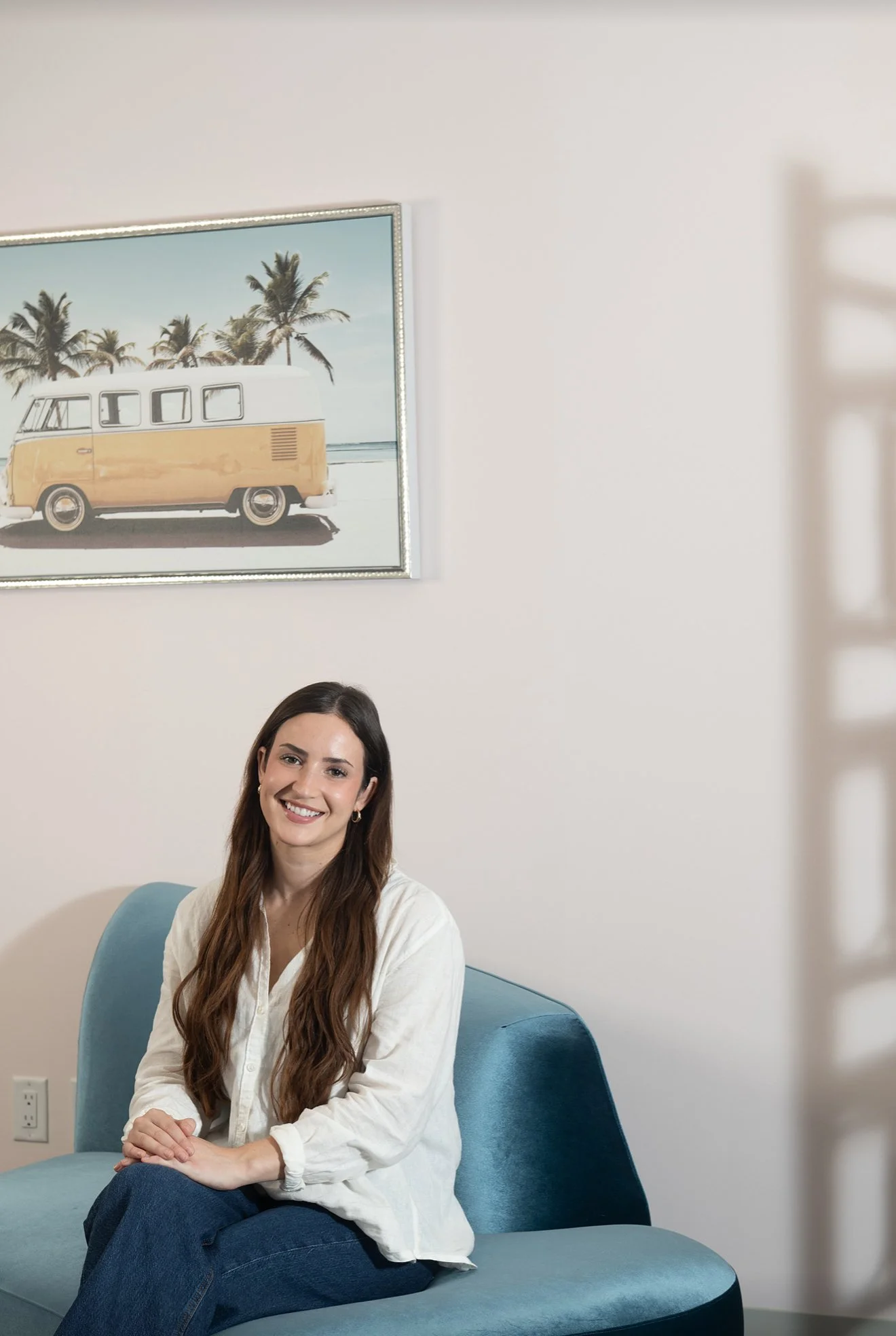 A young woman with long brown hair sitting on a blue couch, smiling, in a room with a framed picture of a vintage yellow and white van with palm trees in the background.