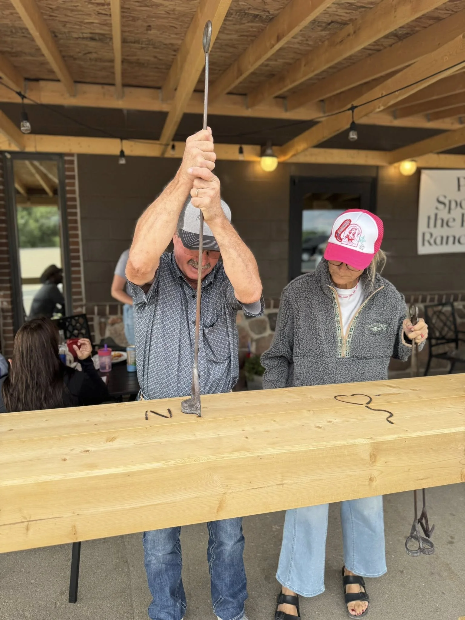 Two people, a man and a woman, are standing behind a large wooden table outside. The man is wearing a gray cap and a short-sleeved shirt, and is holding a large metal spatula upright. The woman is wearing a pink and white cap, a gray fleece jacket, a