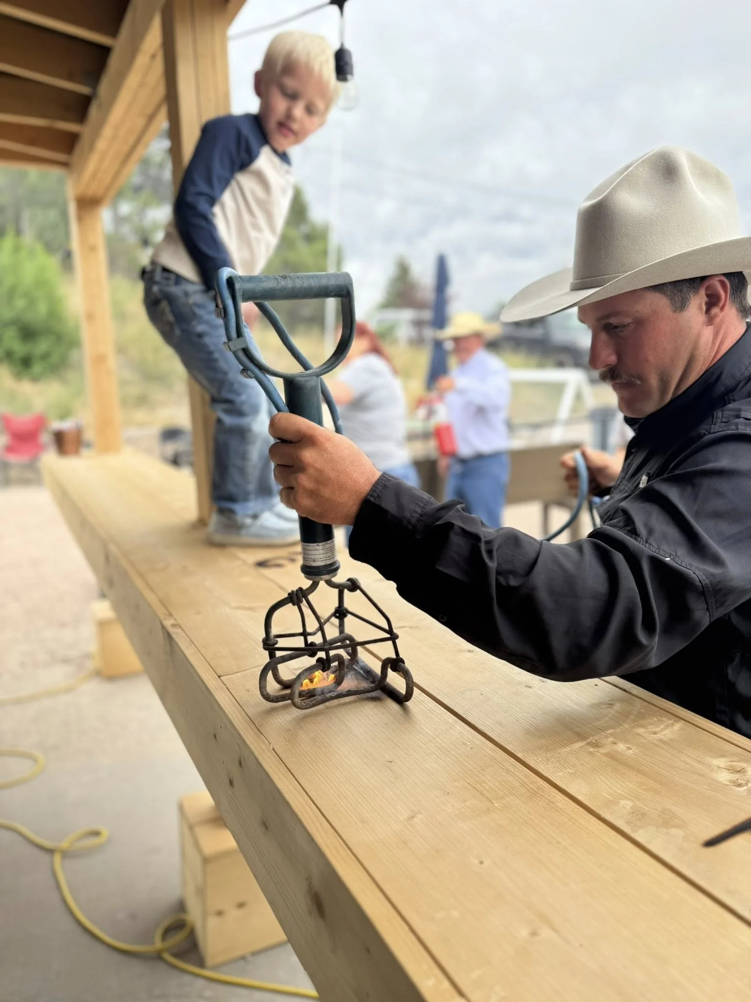 A man in a cowboy hat uses a soldering iron on a piece of wood, with a young boy standing on the wood observing. There are two people in the background also working on a woodworking project.
