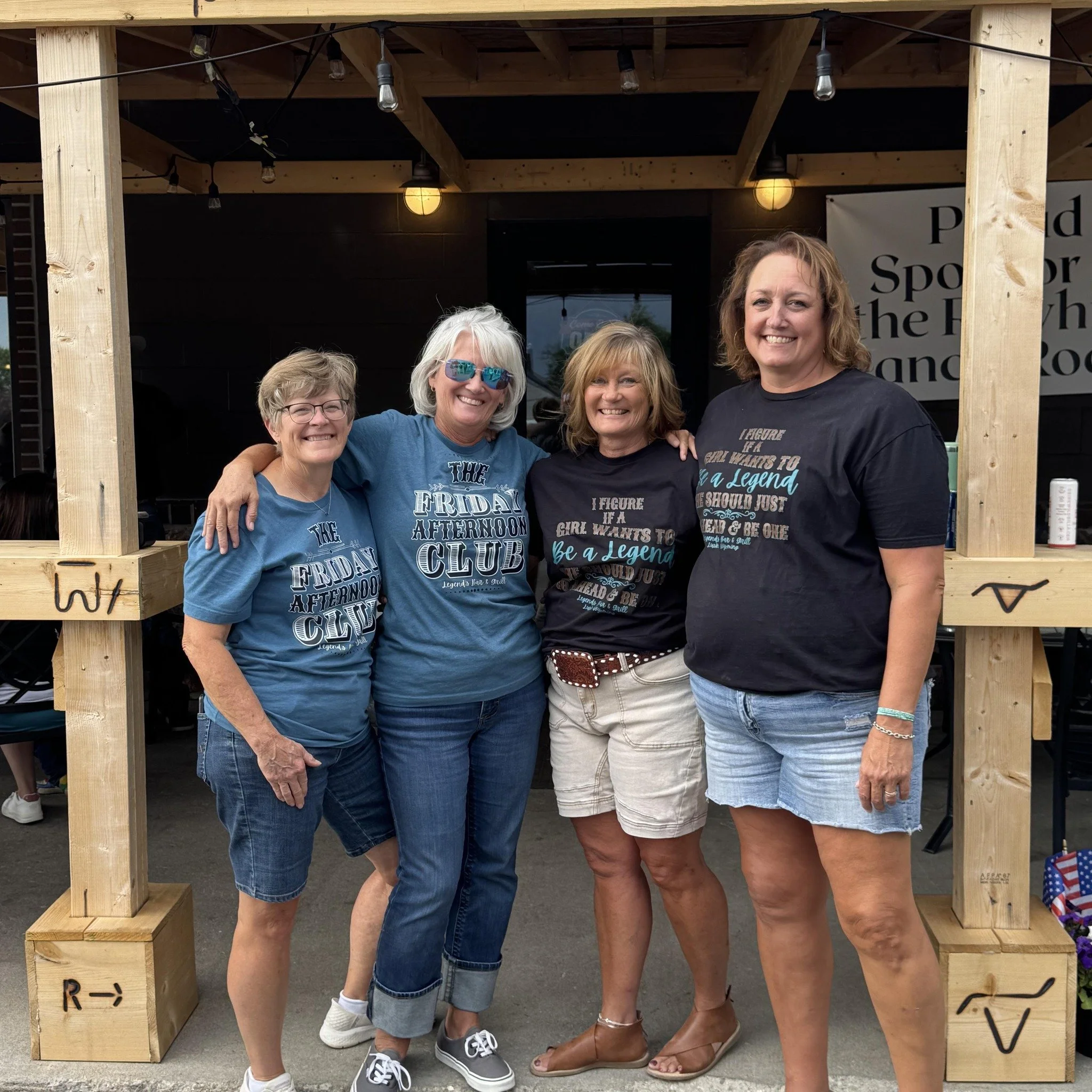 Four women standing together outdoors, smiling, under a wooden arch with string lights, wearing matching T-shirts related to the Friday Afternoon Club.
