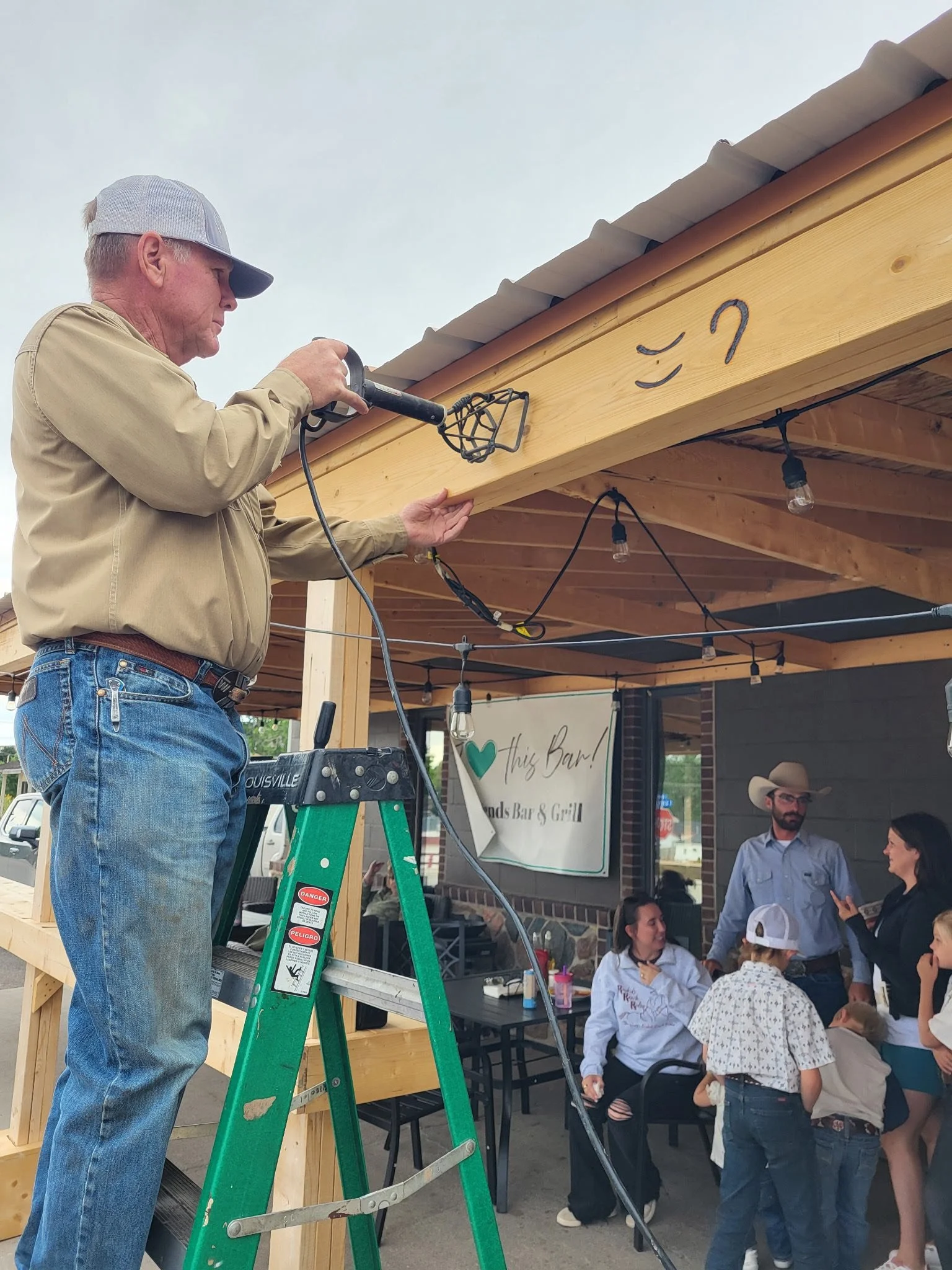 Man on a ladder installing a light fixture under a wooden structure at an outdoor event, with people gathered underneath and a sign for a bar and grill in the background.