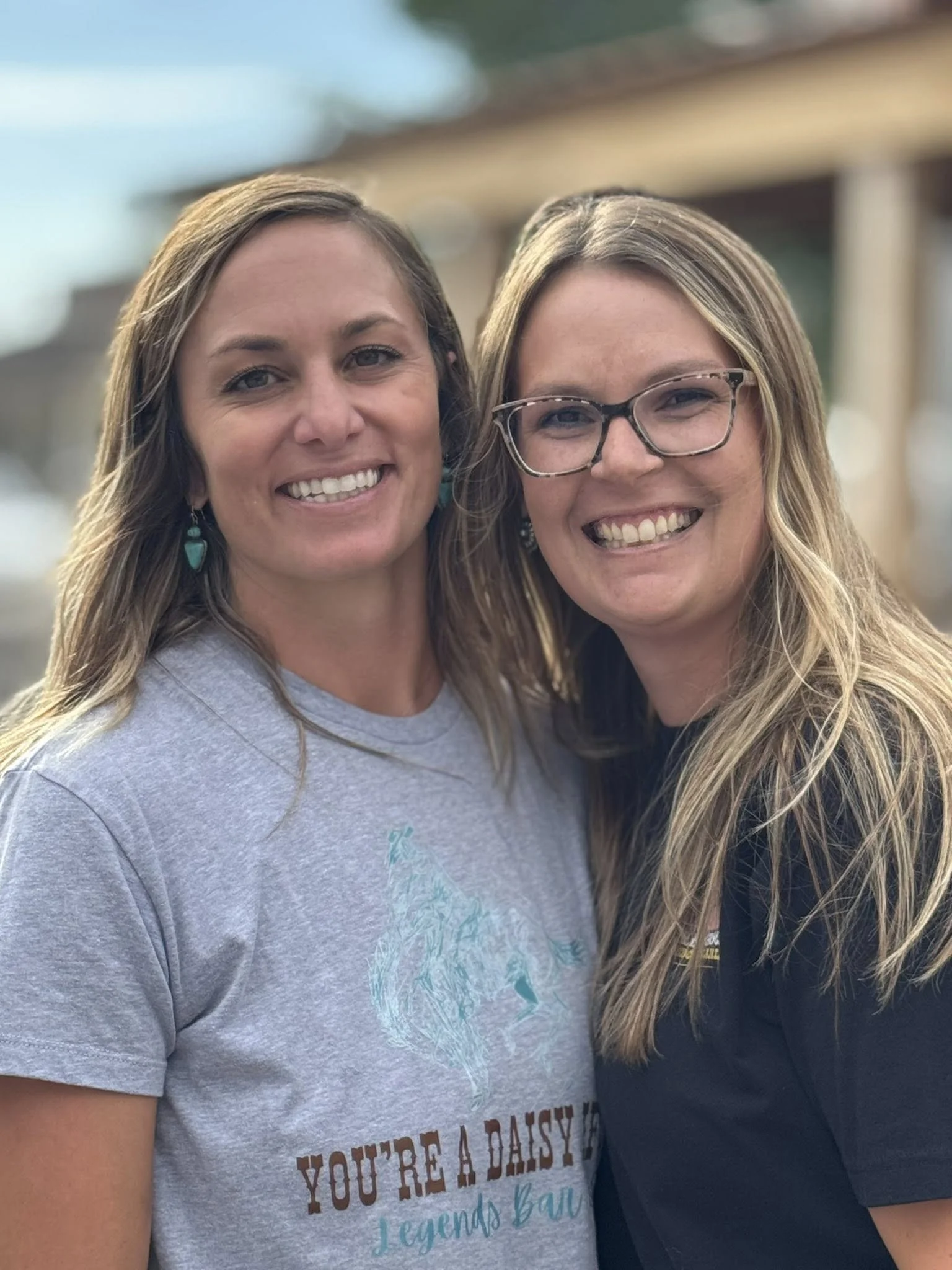 Two smiling women standing close together outdoors with blurred wooden structure in the background. One woman has long brown hair, wearing a gray t-shirt with a print of a horse and the text 'You're a Daisy' and turquoise earrings. The other woman ha