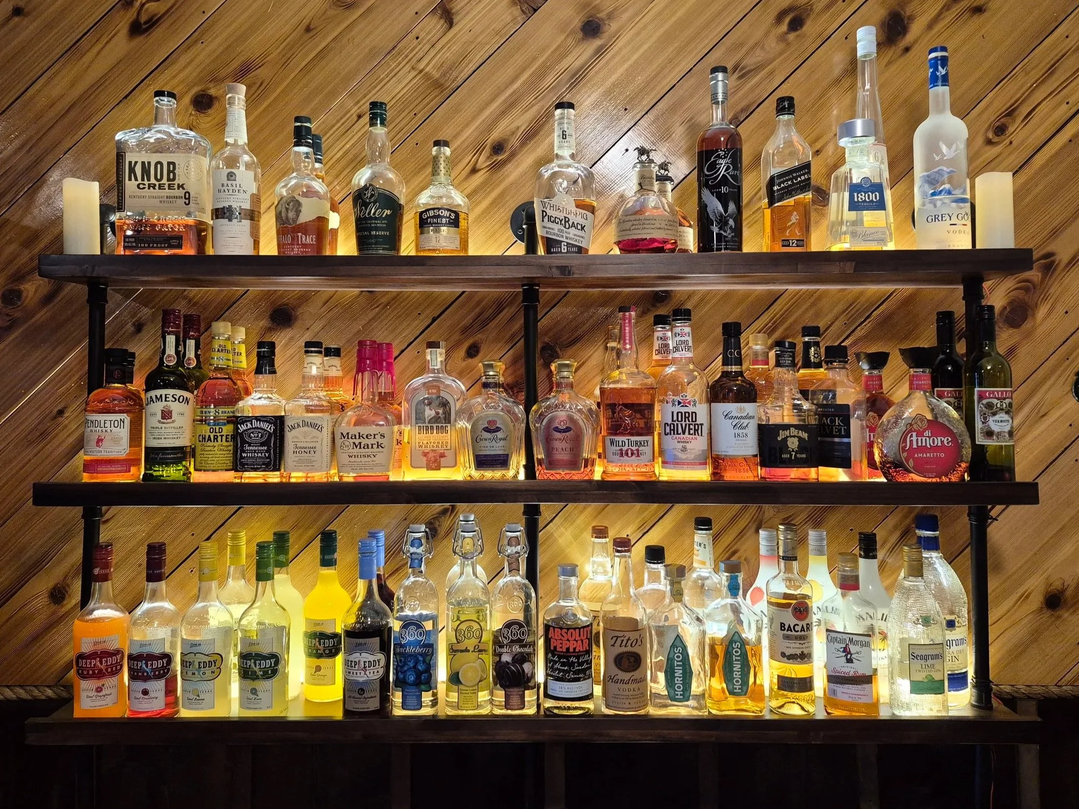 A wooden bar shelf displaying various bottles of whiskey and liquor, illuminated from underneath, against a wood-paneled wall.