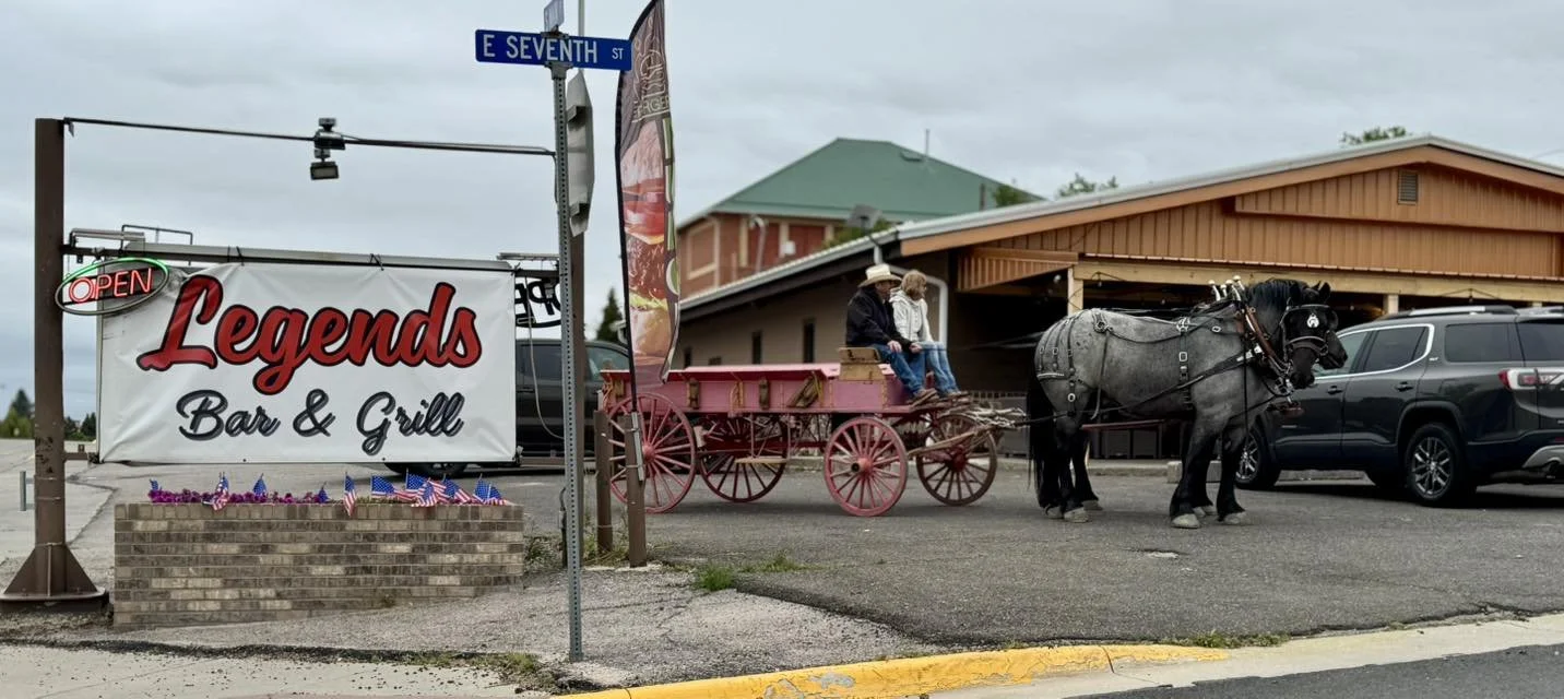 A street corner with a sign for Legends Bar & Grill, an open sign, and small American flags. There is a gray horse with a harness hitched to a pink and brown vintage wagon with two people sitting inside, and a person standing nearby. Parked cars are 