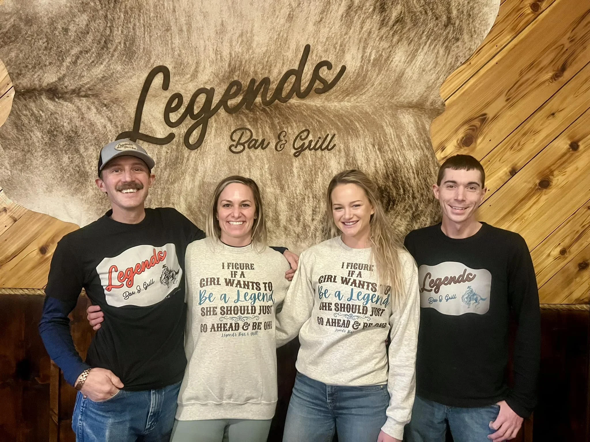 Four smiling people standing in front of a wall with a sign that reads "Legends Bar & Grill." Two women and two men, all wearing casual clothing, smiling and posing for the photo indoors.
