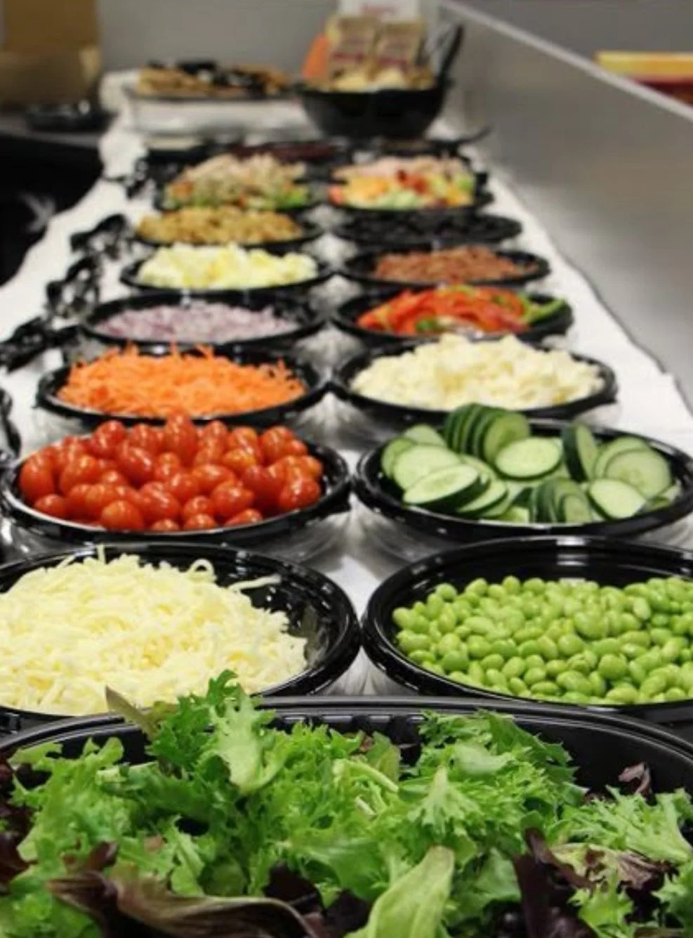 Arrange of salad ingredients in black bowls, including cherry tomatoes, shredded cheese, sliced cucumbers, green peas, shredded lettuce, grated cheese, chopped carrots, chopped onions, diced tomatoes, and other vegetables, set on a white table.