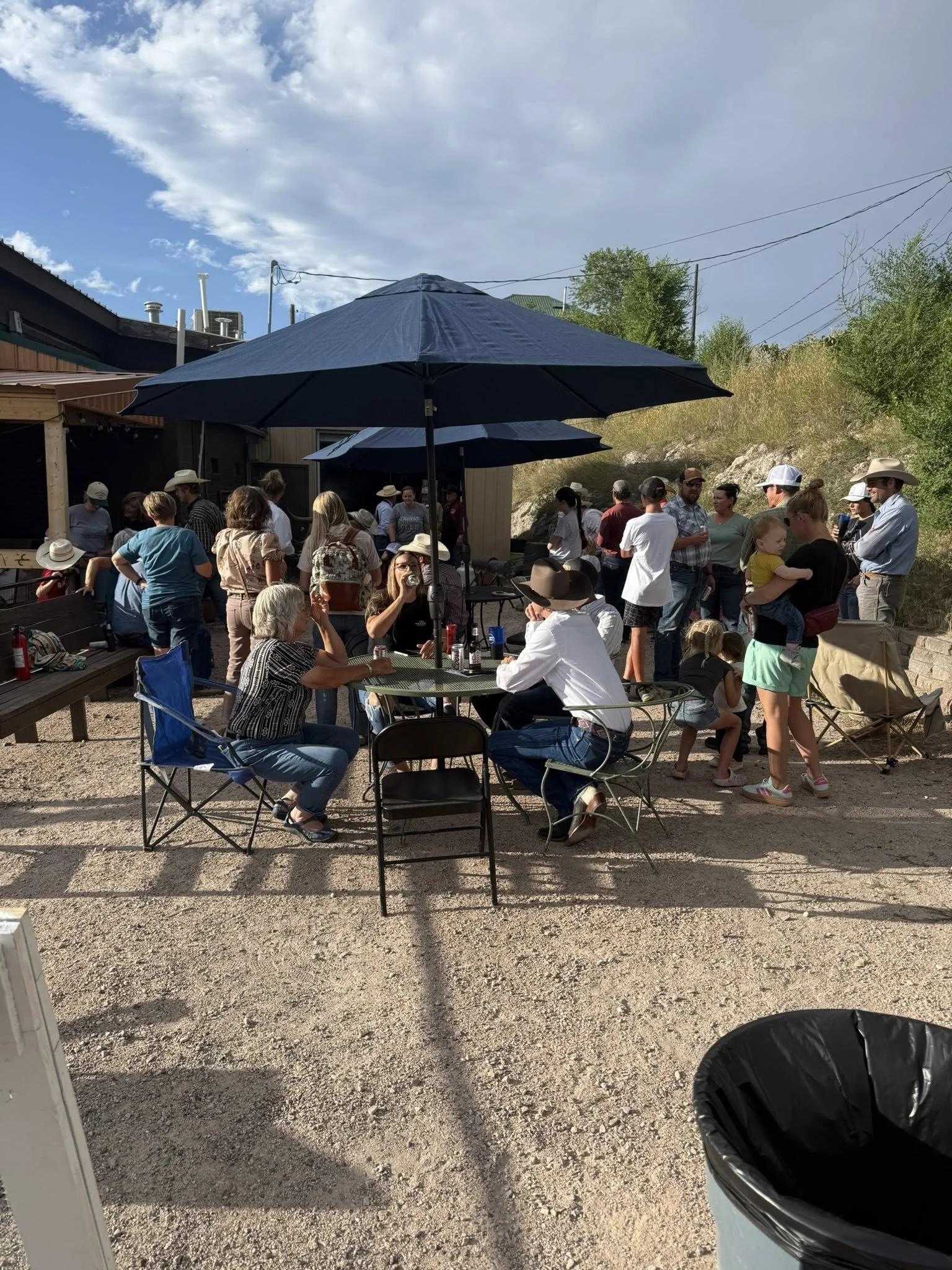 People gathering outdoors, some seated at tables with umbrellas, others standing and socializing, in a casual setting with a building, gravel ground, and a trash can in the foreground.