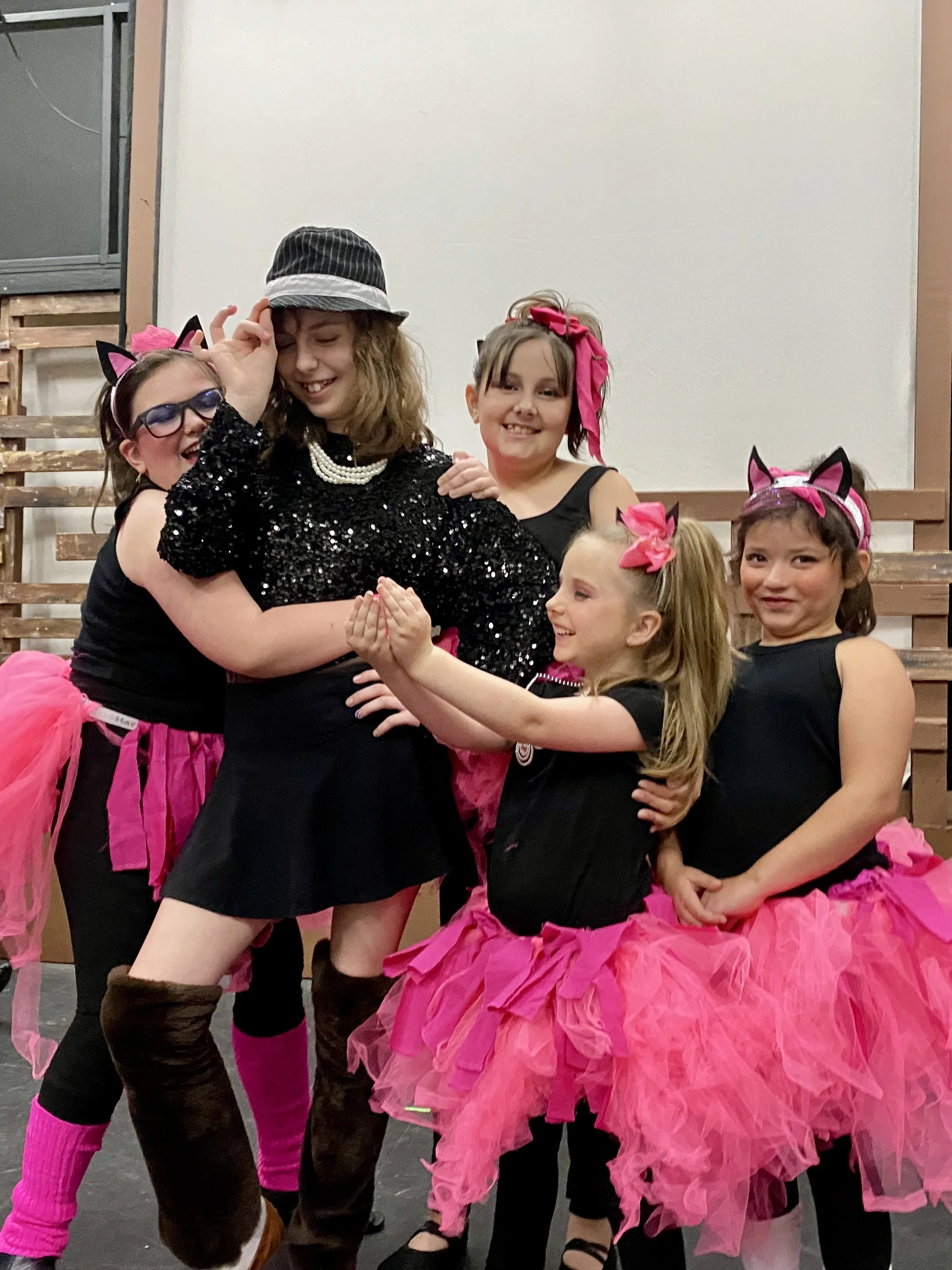Group of young girls dressed in black costumes with pink tutu skirts, some wearing cat ear headbands, and one in a black sequined outfit with a bucket hat, posing playfully in a room with wooden benches and a plain white wall.