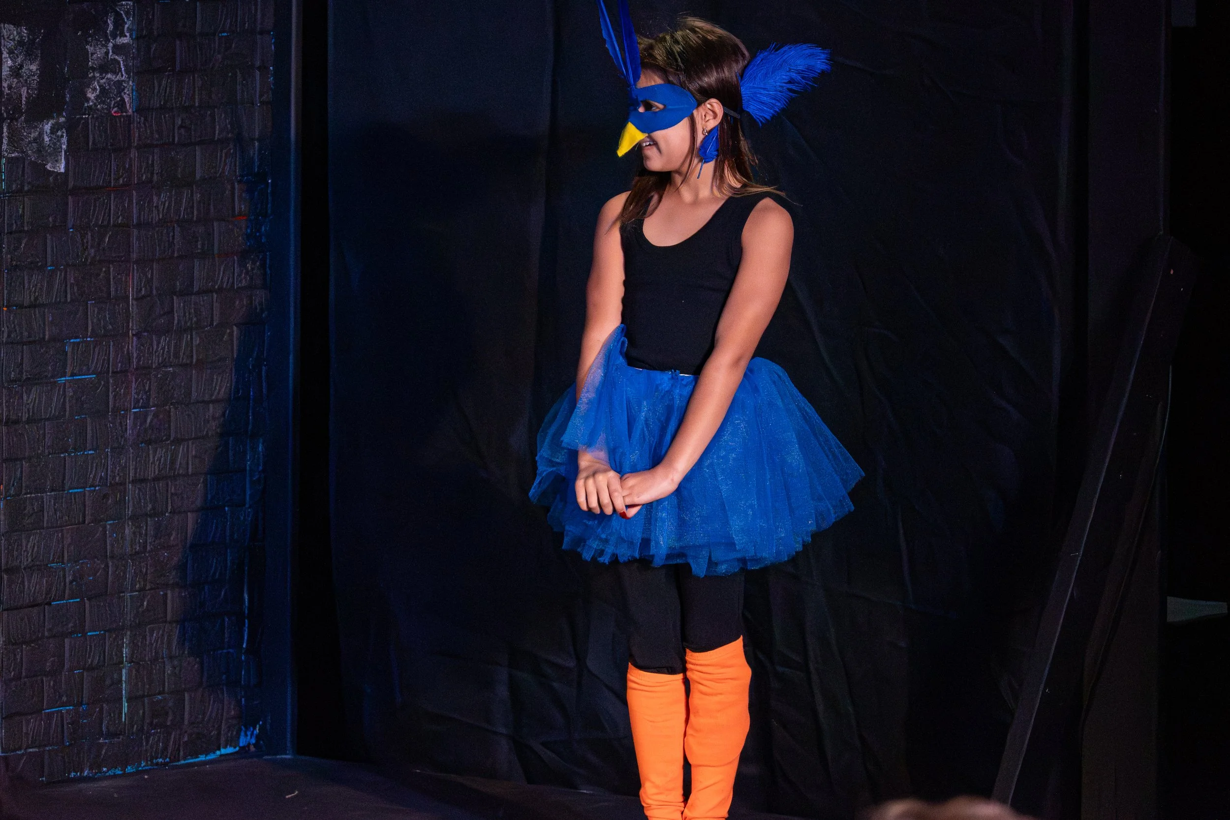A young girl wearing a blue bird mask with a yellow beak and matching blue feathered headpiece, dressed in a black top and a blue tutu, stands in a dark setting with black curtains and a brick wall, smiling and holding her hands together.