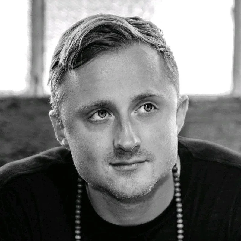 Black and white portrait of a young man with short, slicked-back hair, light-colored eyes, and a slight smile, wearing a beaded necklace and a dark shirt.