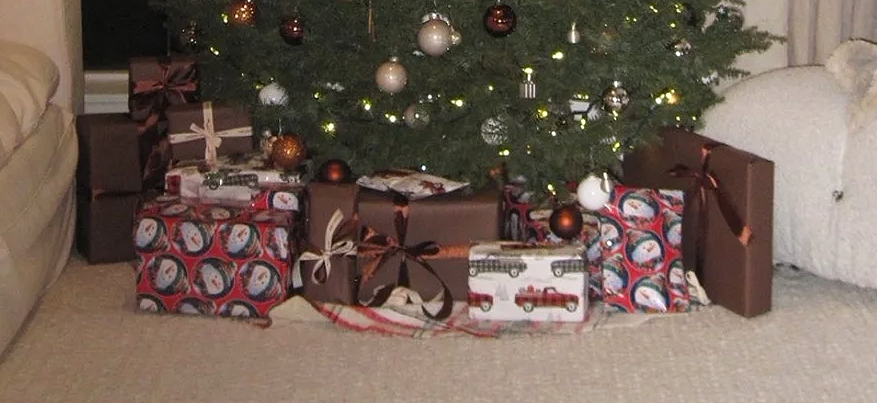 Christmas tree with ornaments and string lights, surrounded by wrapped gifts with various patterns and ribbons on a carpeted floor.