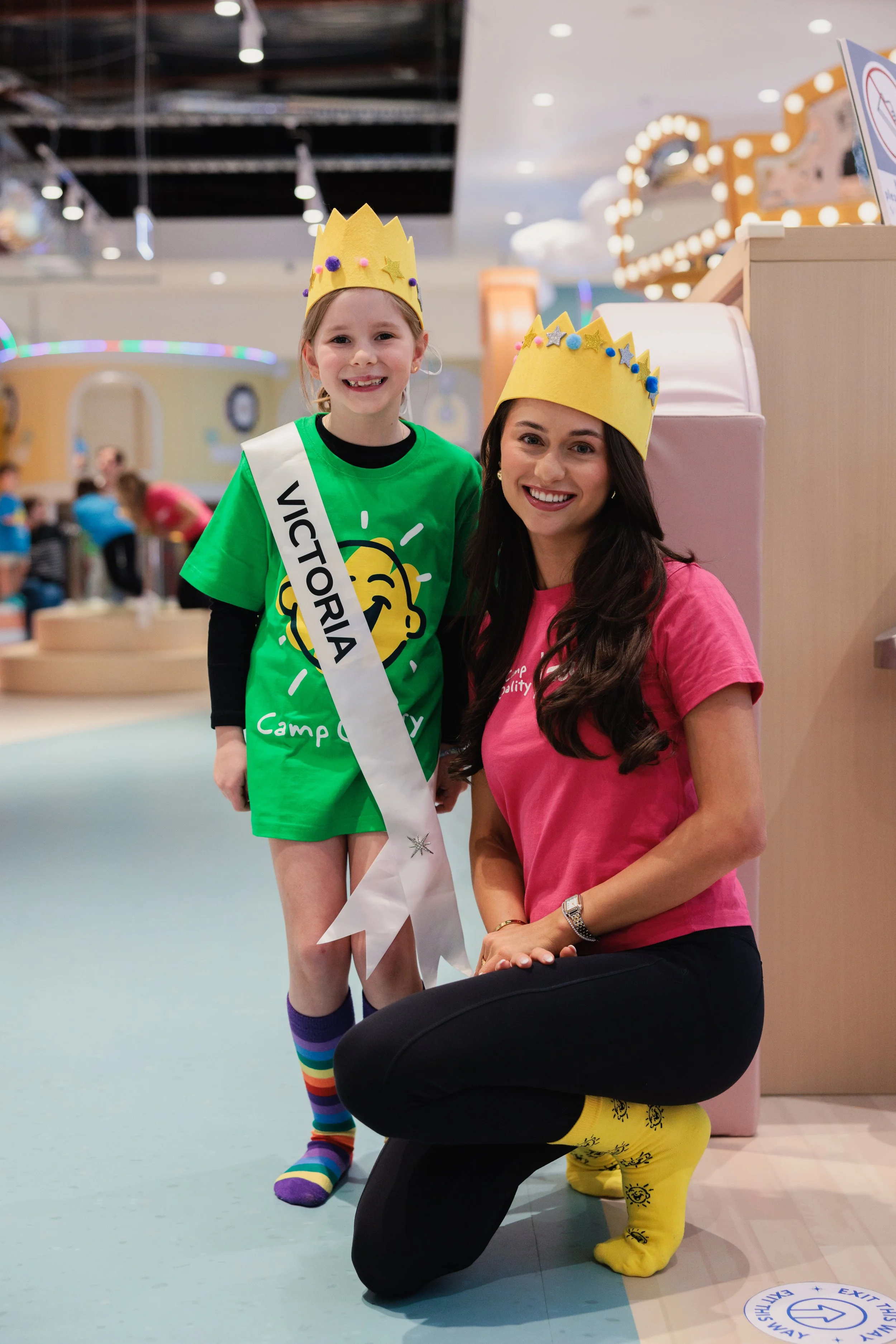 Young girl and woman wearing yellow paper crowns and bright clothing, smiling for the camera at an indoor play area or amusement venue.