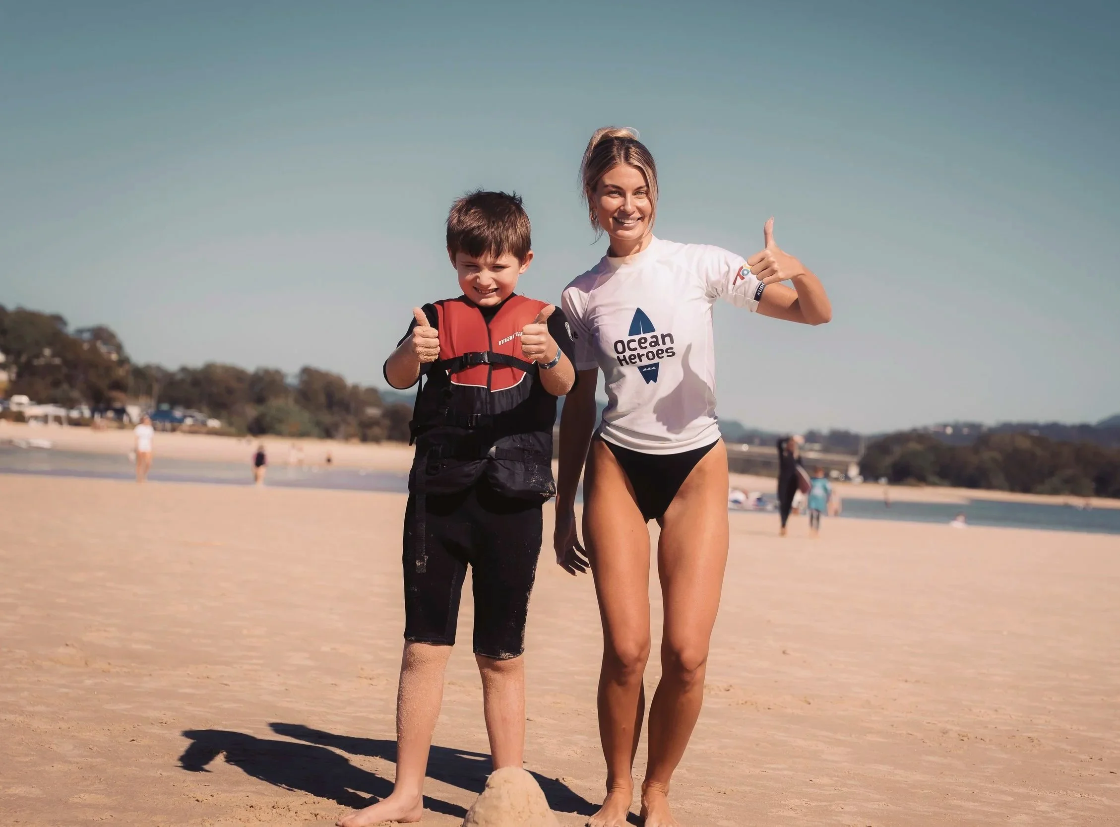A young boy and a woman at the beach, both smiling and giving thumbs up. The boy is wearing a life vest and black swim shorts, while the woman is wearing a white t-shirt with the text 'Ocean Heroes' and black bikini bottoms. They are standing on the sand with a sandcastle in front of them.