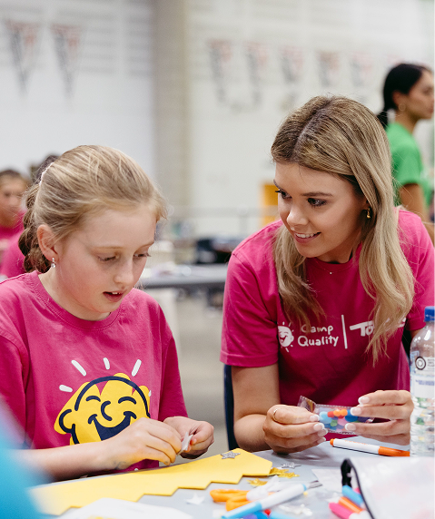 Two girls in pink camp shirts working together at a table with craft supplies inside a busy classroom or camp setting.