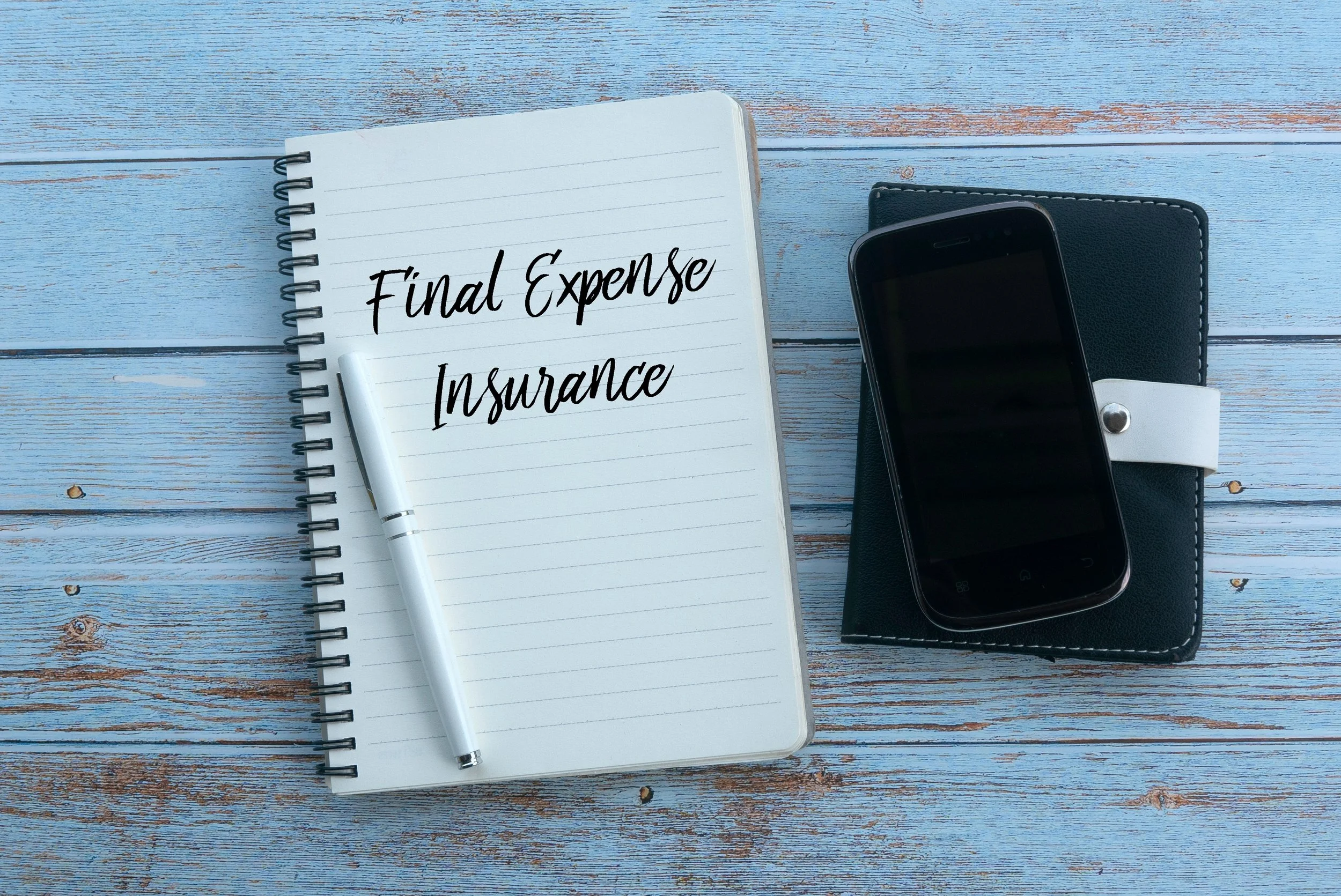Notebook with 'Final Expense Insurance' written on it, pen, and a smartphone with a black cover on a blue wooden table.