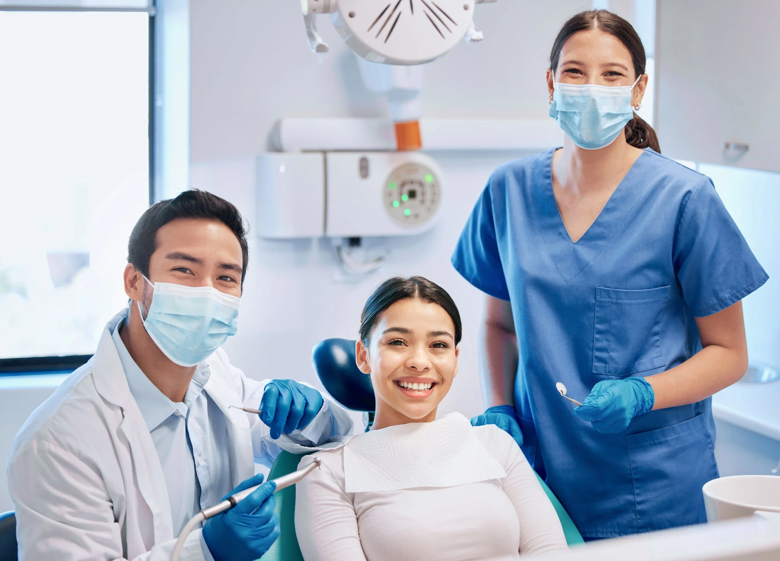 A smiling woman patient sitting in a dental chair, accompanied by a male dentist and a female dental assistant, all wearing face masks and gloves in a dental clinic.