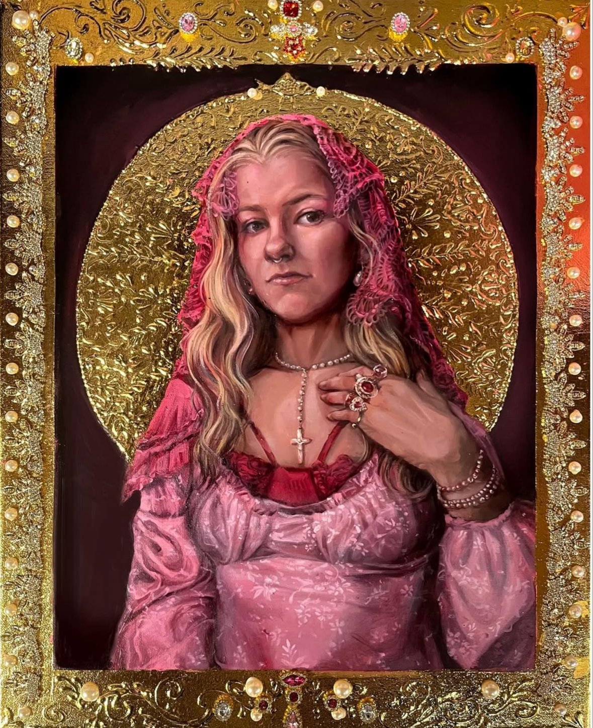 A woman with long wavy blonde hair dressed in pink, wearing jewelry including rings, bracelets, a pearl necklace, and a cross pendant, posing in front of a decorative background with gold and red accents.