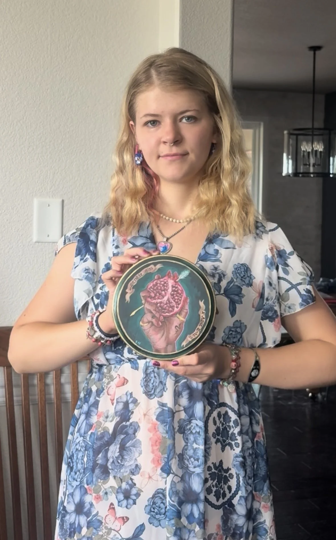 Young woman standing indoors, holding a decorative plate with a pomegranate illustration. She has blonde, wavy hair, wears colorful jewelry and a floral dress.